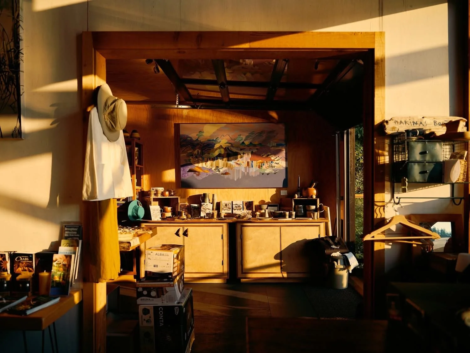 Interior of a store with wooden furniture, pottery, and artwork on the wall, illuminated by warm sunlight.