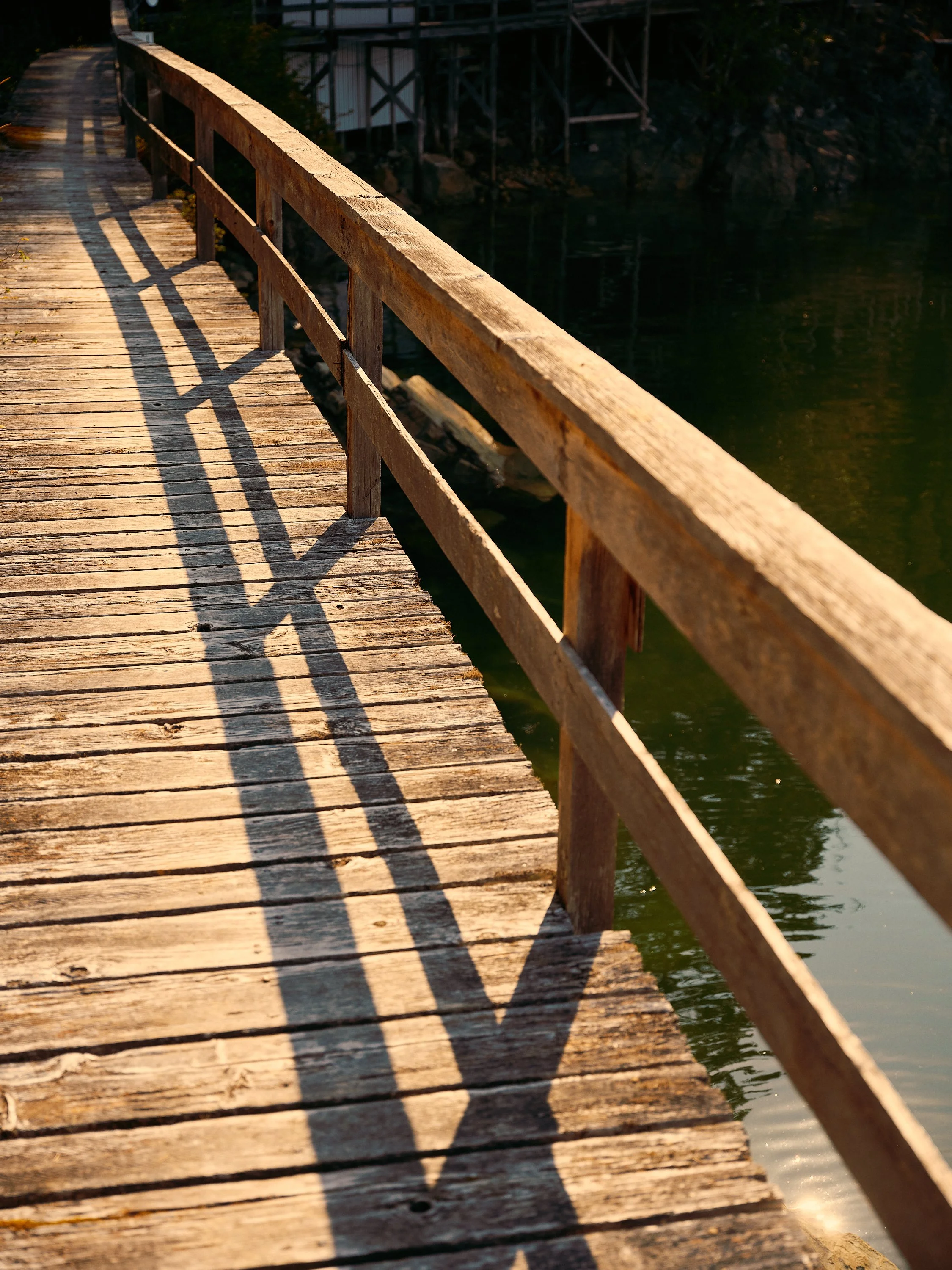 A weathered wooden bridge casting shadows over the water below, with buildings on stilts in the background.