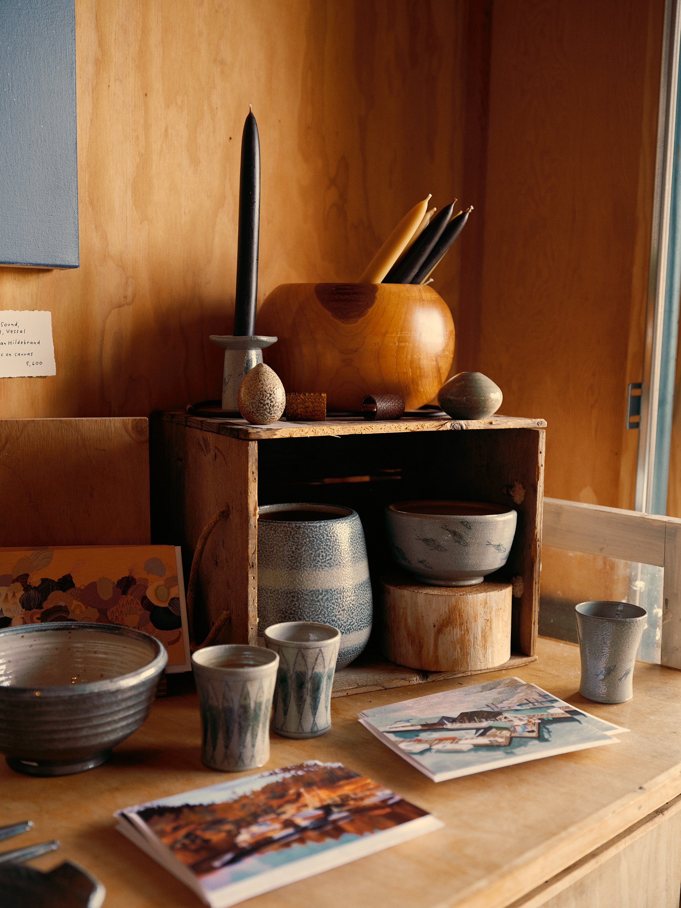 Display of ceramic pottery and pottery-related items, including bowls, cups, and vases, arranged on a wooden shelf and table, with a wooden wall background.