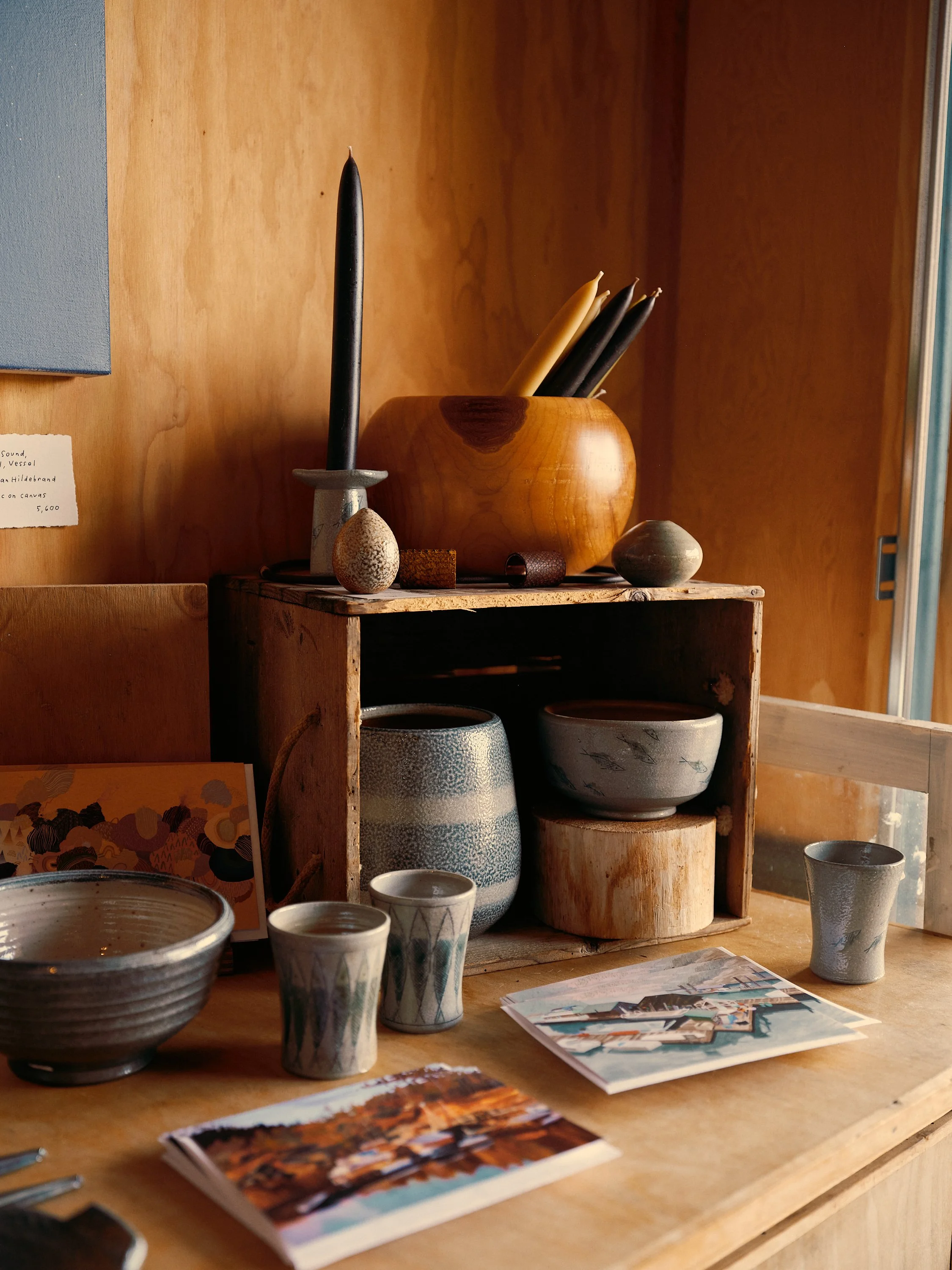 Display of pottery items including bowls, cups, and vases arranged on a wooden table and shelf, with a wooden wall background.
