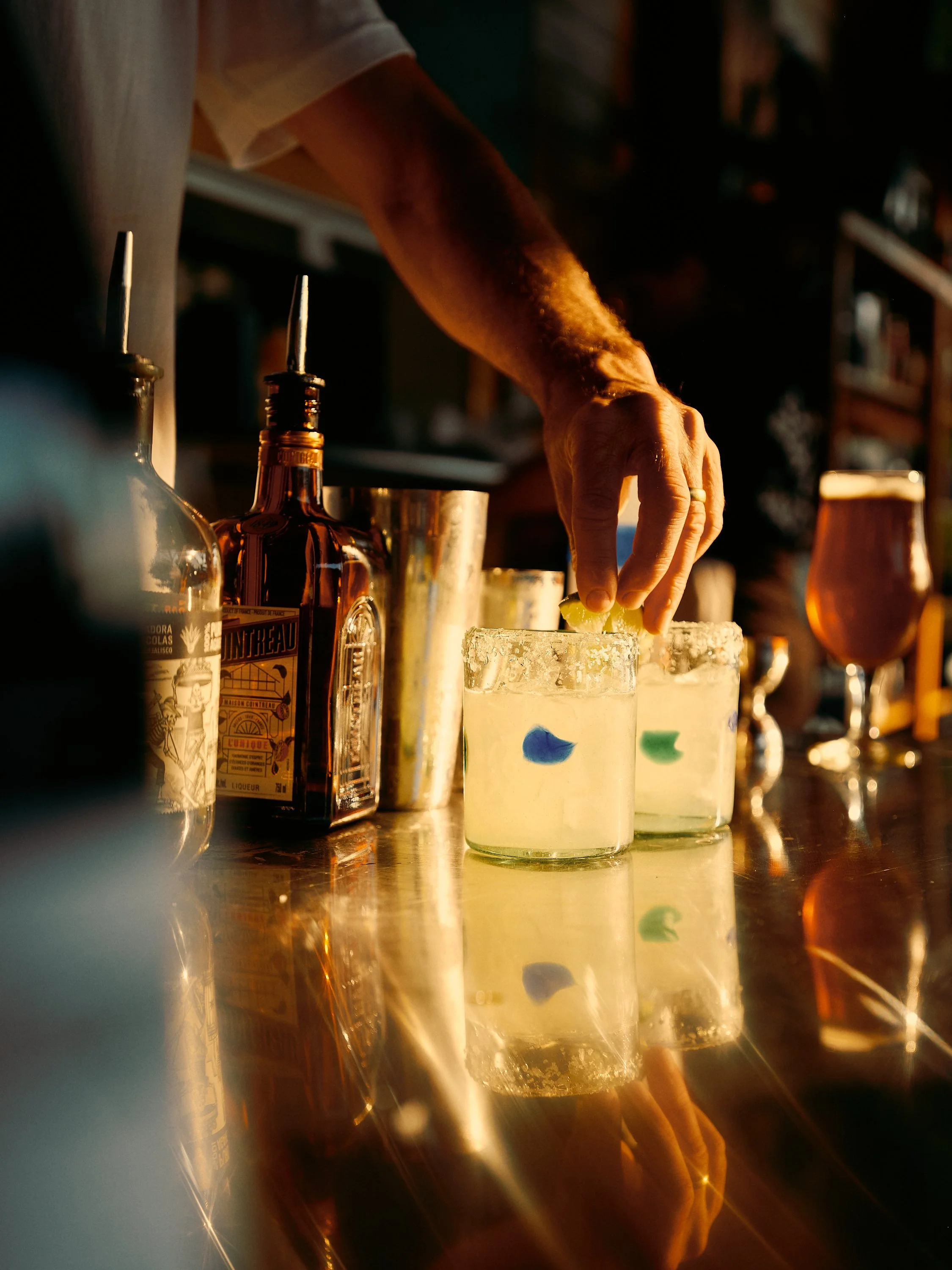 Close-up of a bartender's hand preparing a cocktail at a bar, with bottles and glasses visible on the counter, reflecting warm lighting.