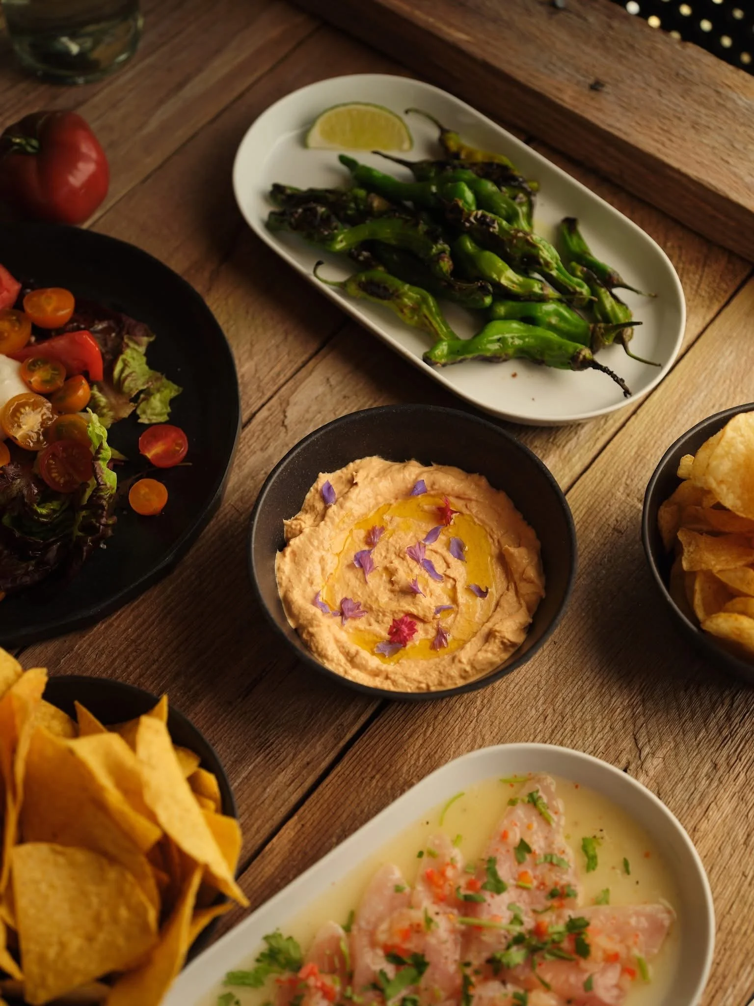 A table with various Mexican dishes including grilled jalapeño peppers with lemon wedge, a bowl of ceviche topped with chopped herbs, a dish of hummus garnished with edible flower petals and olive oil, a bowl of chips, and a salad with cherry tomatoes and lettuce.