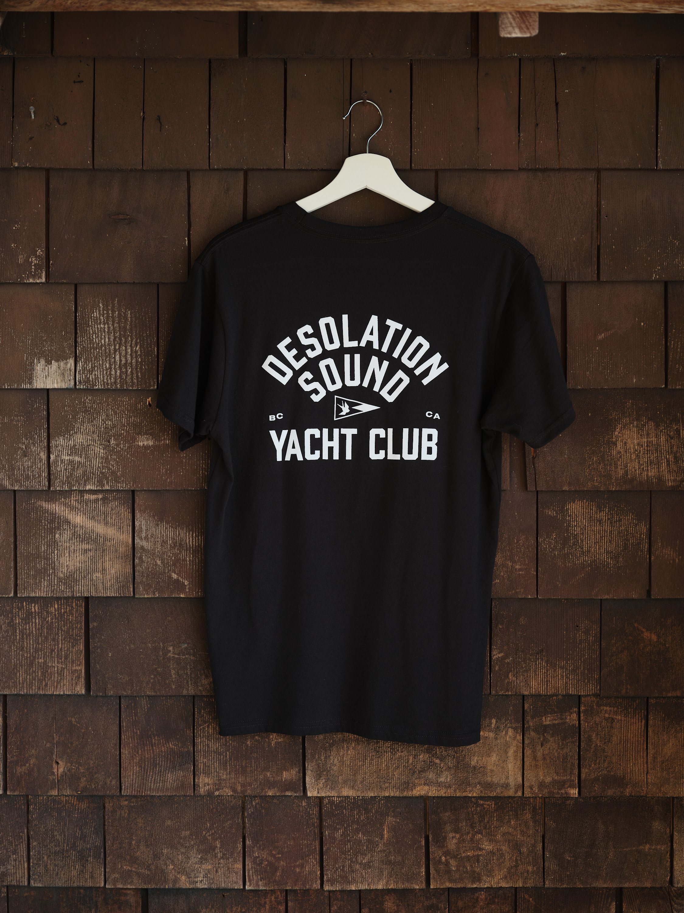 Black t-shirt hanging on a white hanger against a wood shingle wall. The t-shirt has white text that reads 'Desolation Sound Yacht Club' with a small logo and the initials 'BC' and 'CA'.