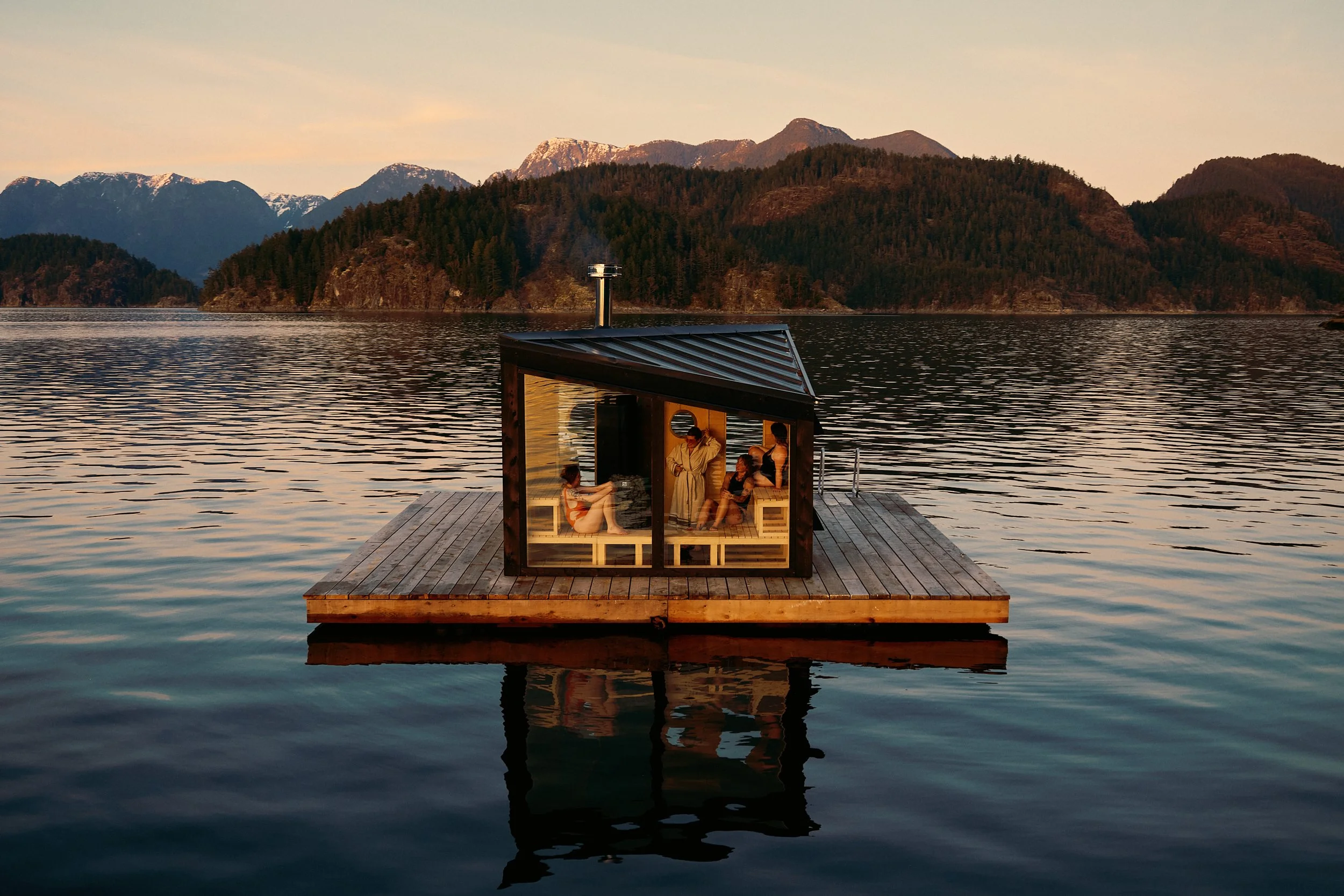 A small wooden floating dock on a body of water with a glass cabin containing people relaxing inside, backdropped by forested hills and mountains at sunset.