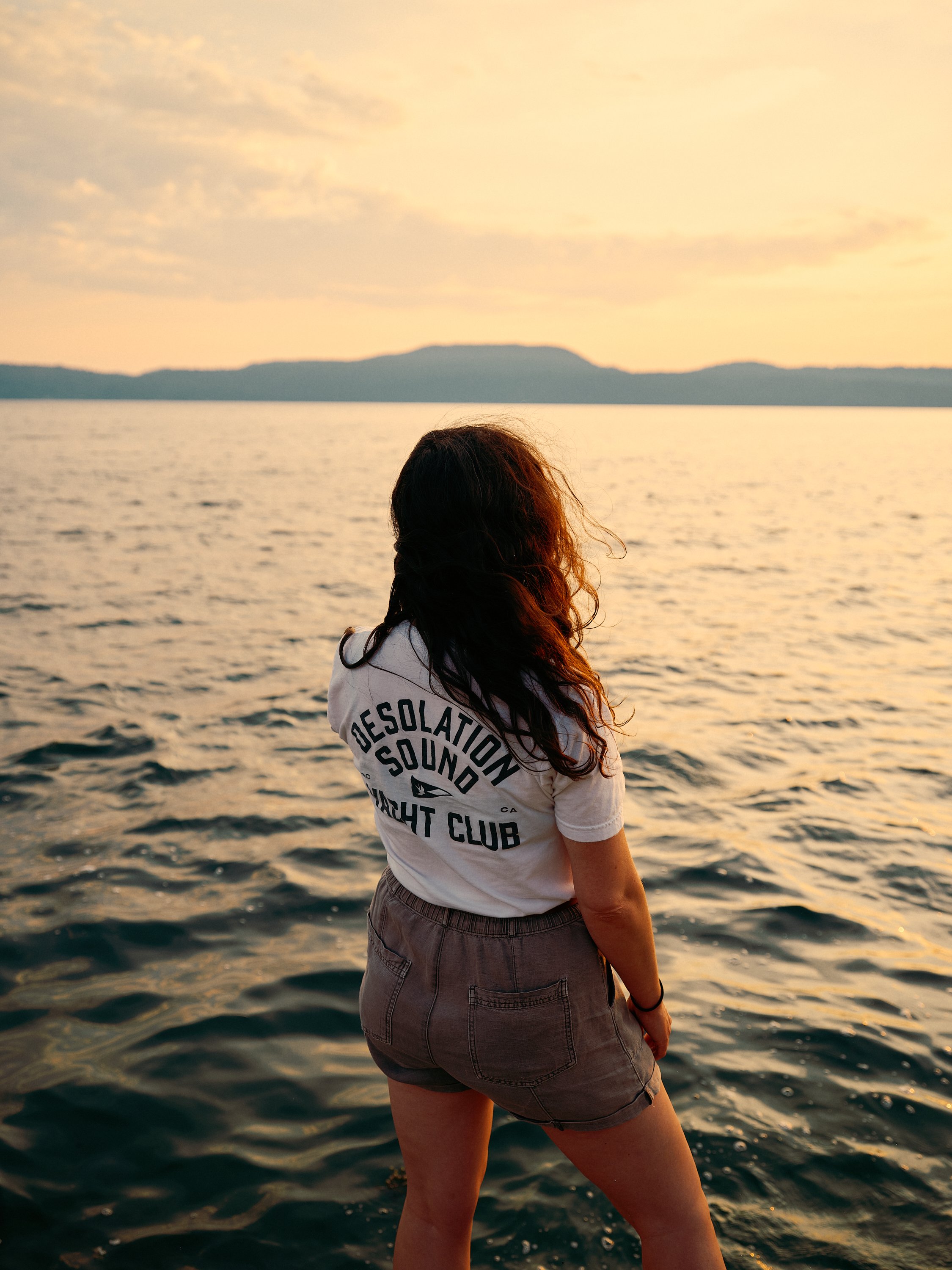 A person with long, dark hair wearing a white t-shirt and shorts standing by a body of water during sunset, looking at distant mountains.
