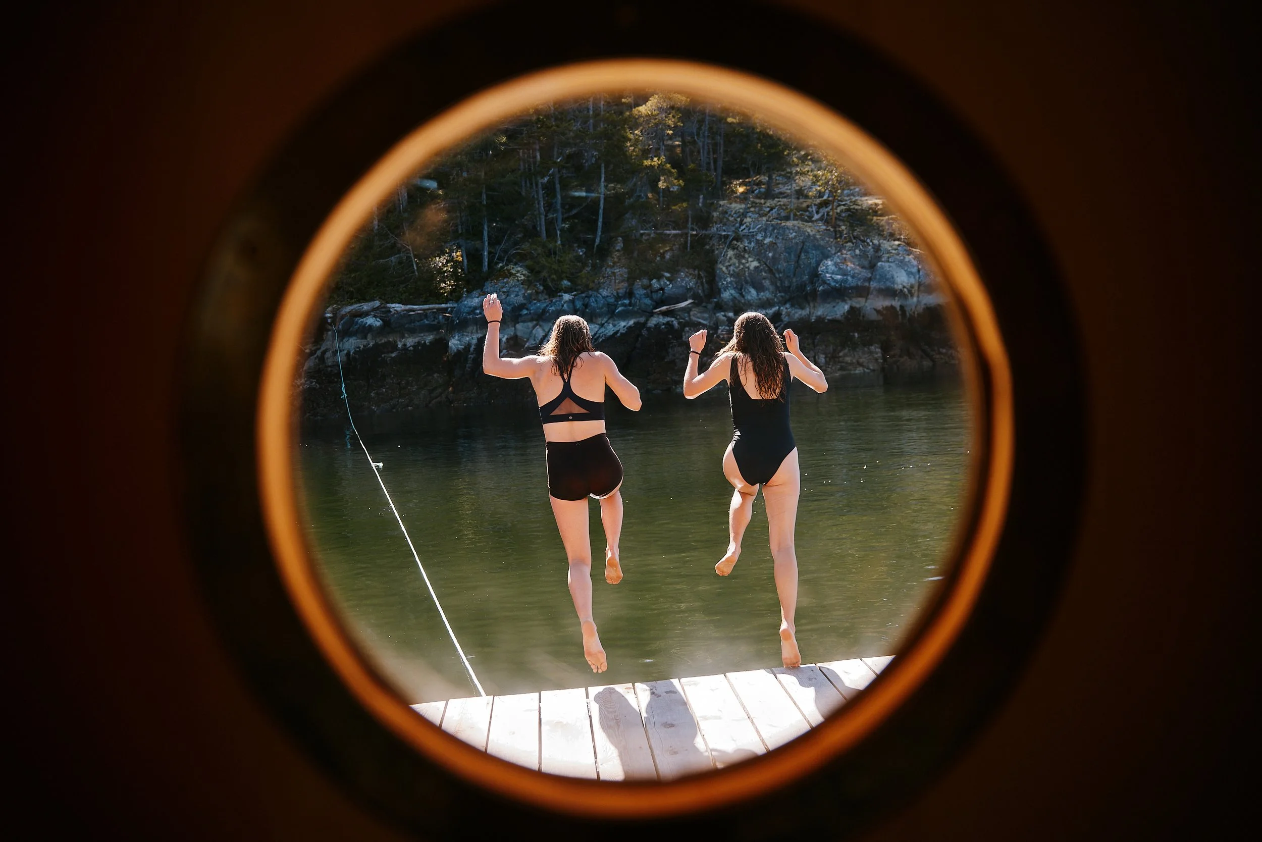 Two women in black swimsuits jumping into the ocean, viewed through a porthole window opening.