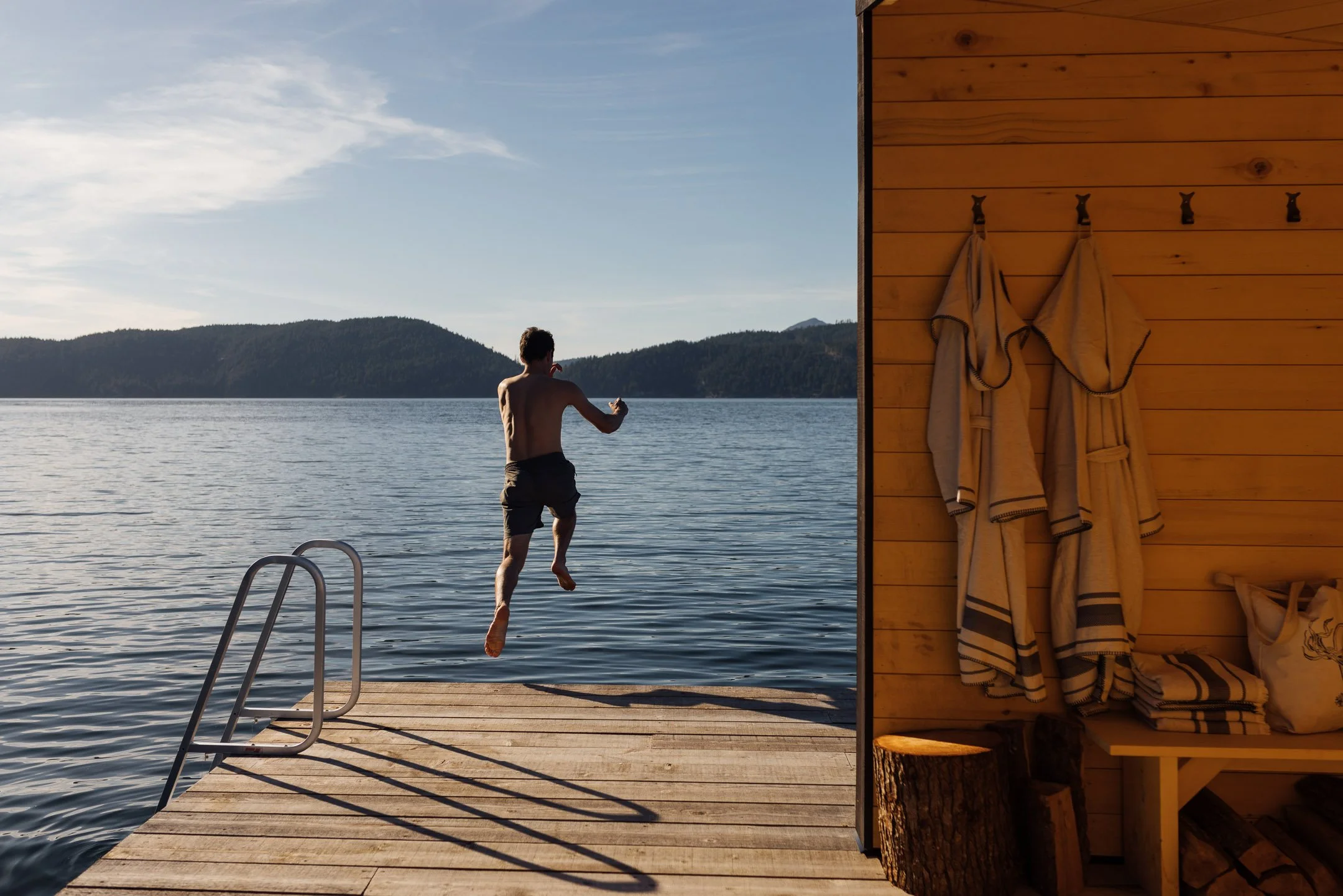 A man in swim trunks jumping into the ocean from a wooden dock, with a mountain landscape in the background. To the right, there is a wooden wall with hooks holding towels and robes.
