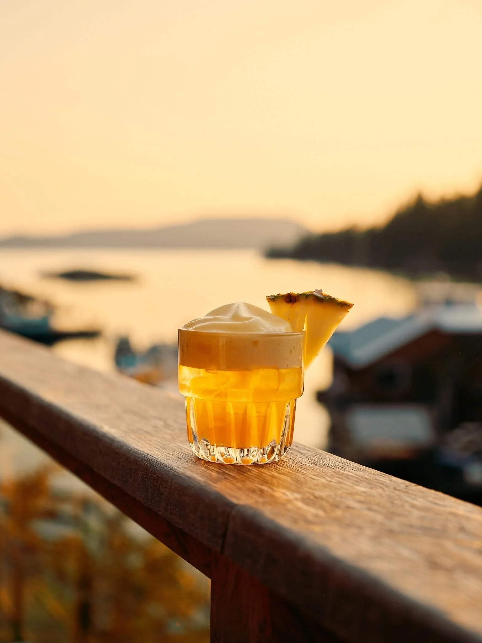 Glass of tropical cocktail garnished with pineapple slice on wooden railing during sunset.