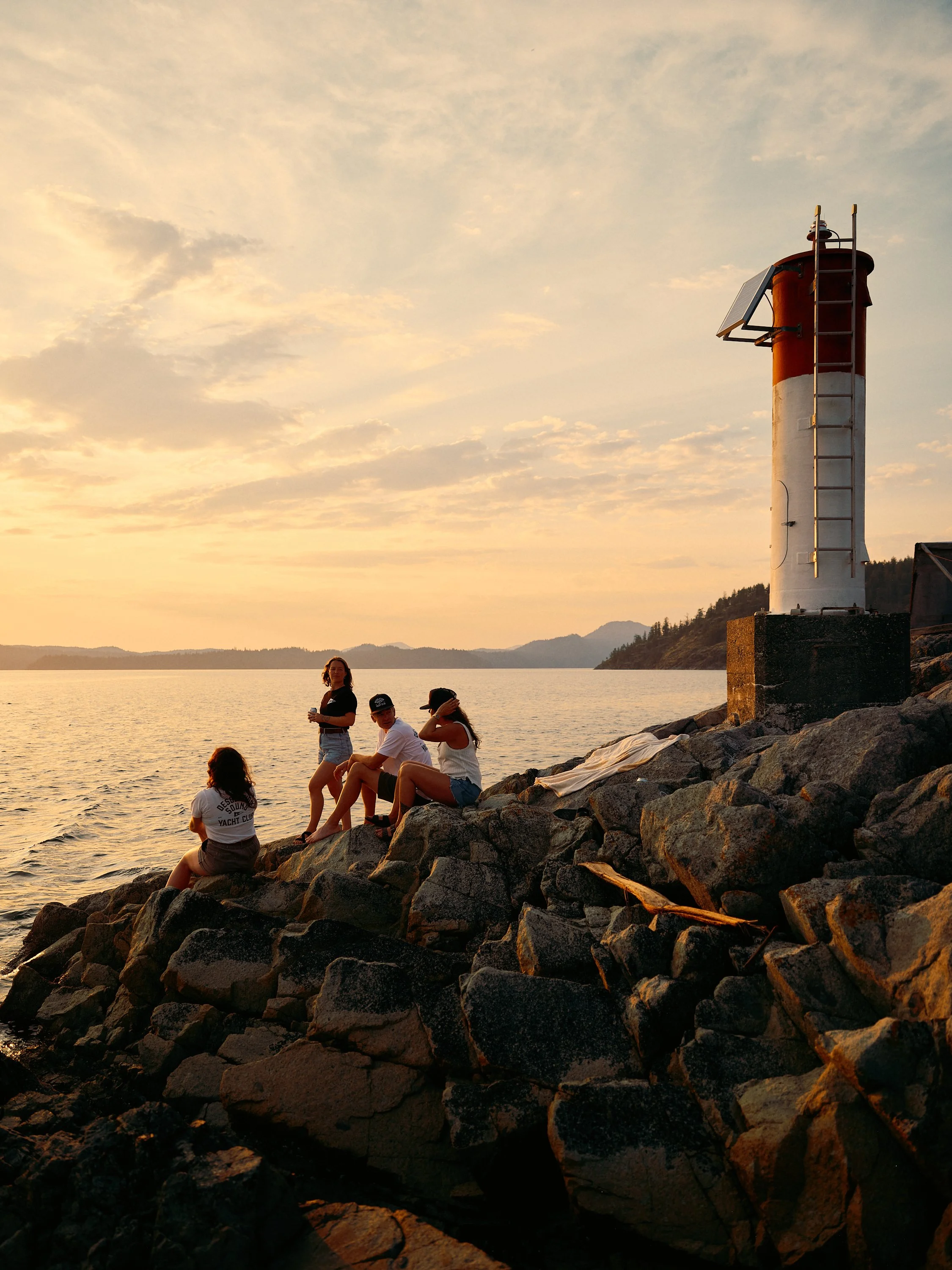 Group of people sitting on rocks by the water during sunset, with a lighthouse to the right and hills in the background.