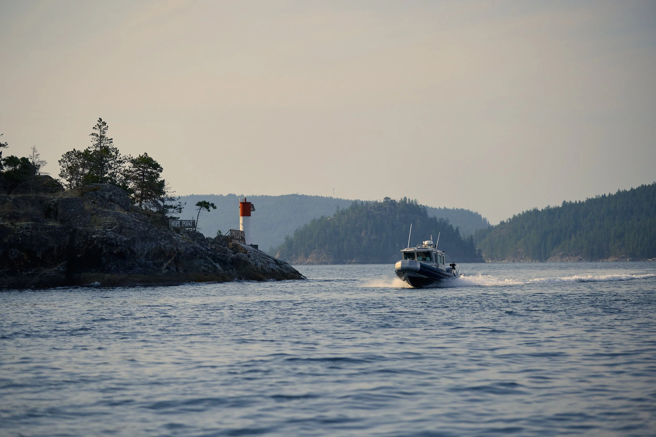 A motorboat speeding through a body of water near rocky shoreline with trees and forested hills in the background, under a cloudy sky.