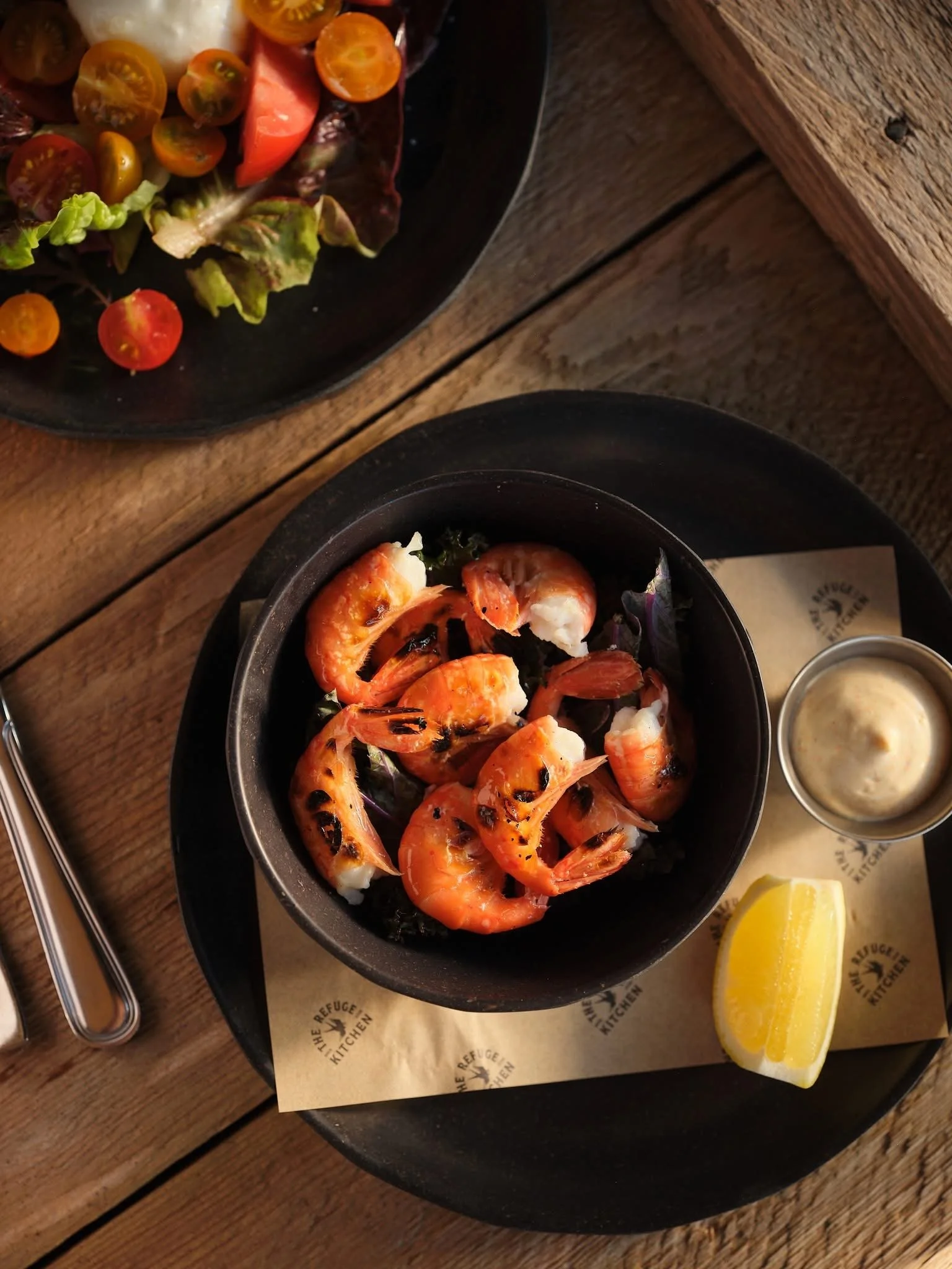 A table with two black bowls containing grilled shrimp and a salad with cherry tomatoes, lettuce, and other vegetables. There is a lemon wedge and a small container of dipping sauce nearby.