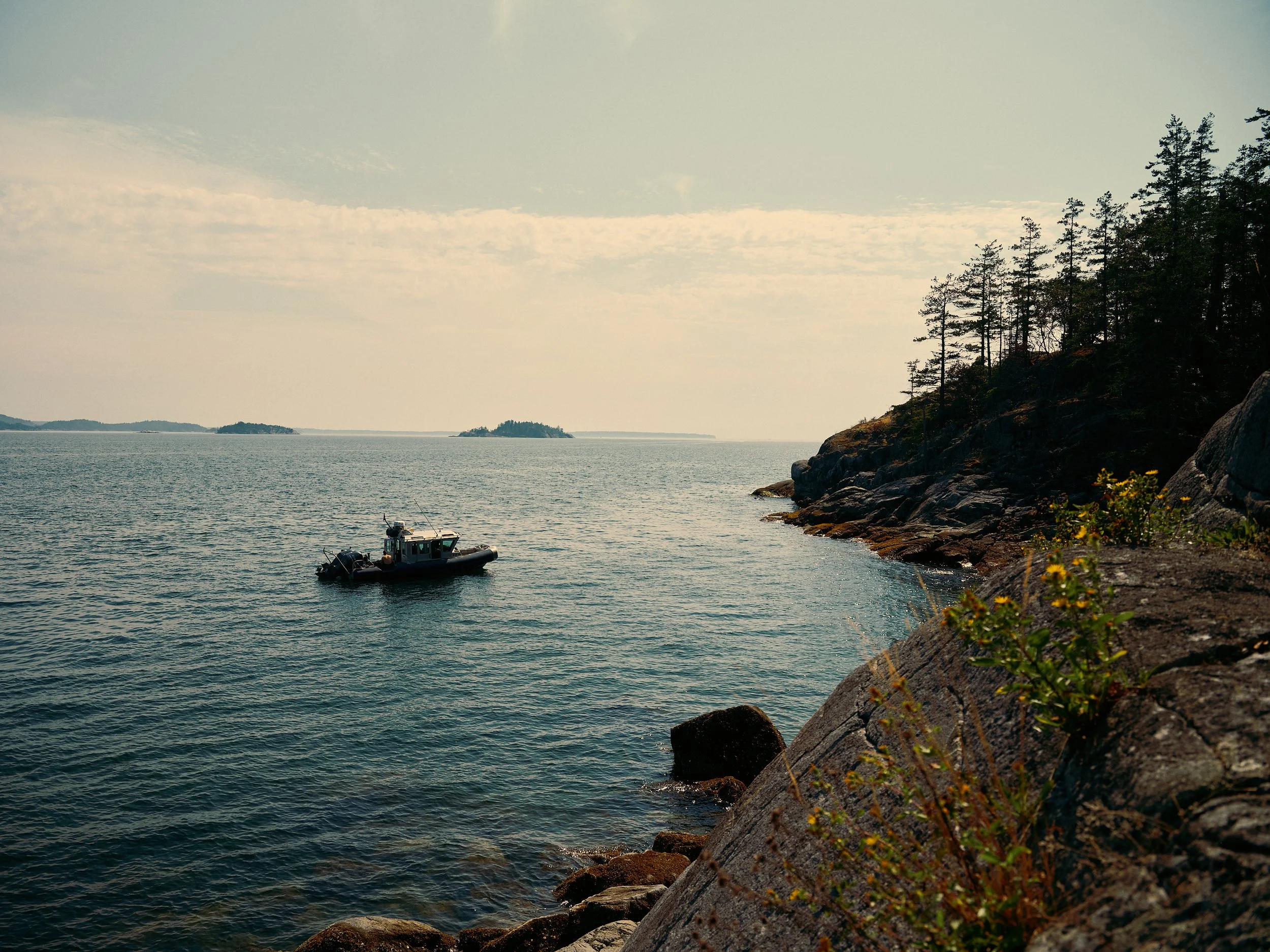 Scenic view of a rocky shoreline with a small boat in the water, and distant islands under a partly cloudy sky.