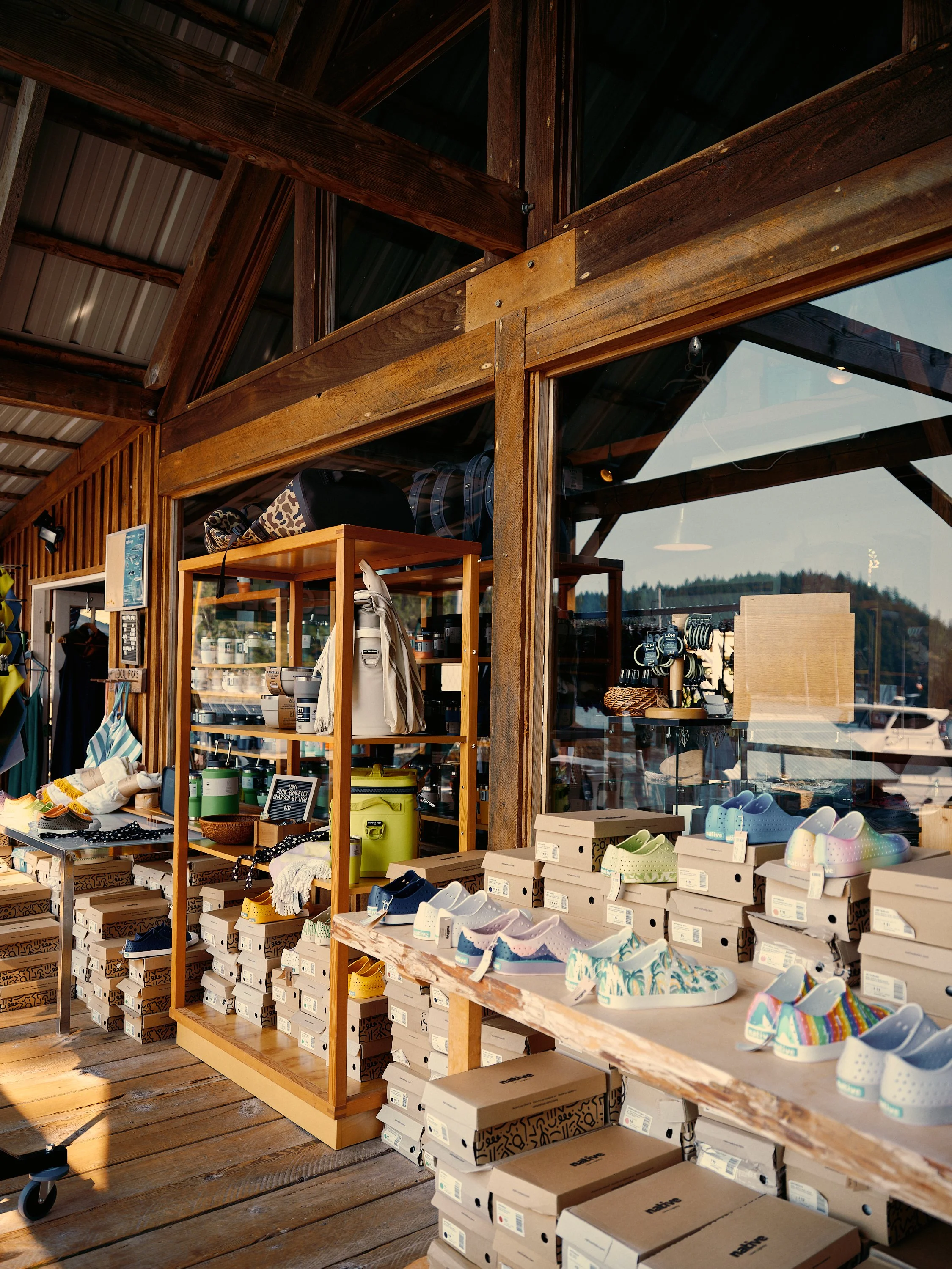 Inside of a retail store selling shoes and outdoor gear, with shoes displayed on wooden tables and shelves, and large windows showing outdoor scenery.