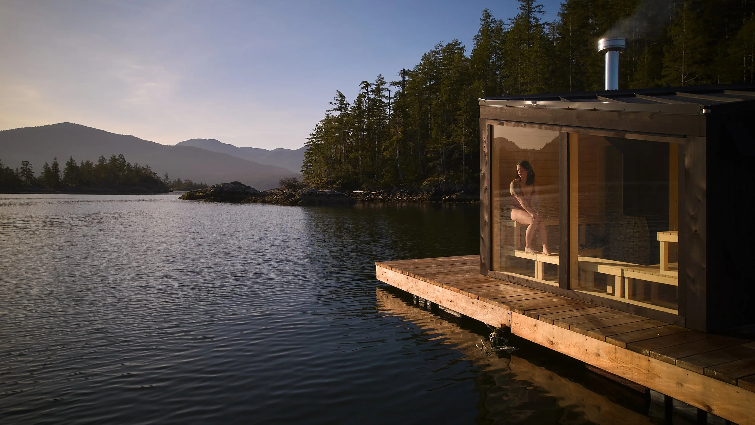 A person sitting on a wooden bench inside a glass sauna overlooking a calm waters, surrounded by forested mountains during sunset.