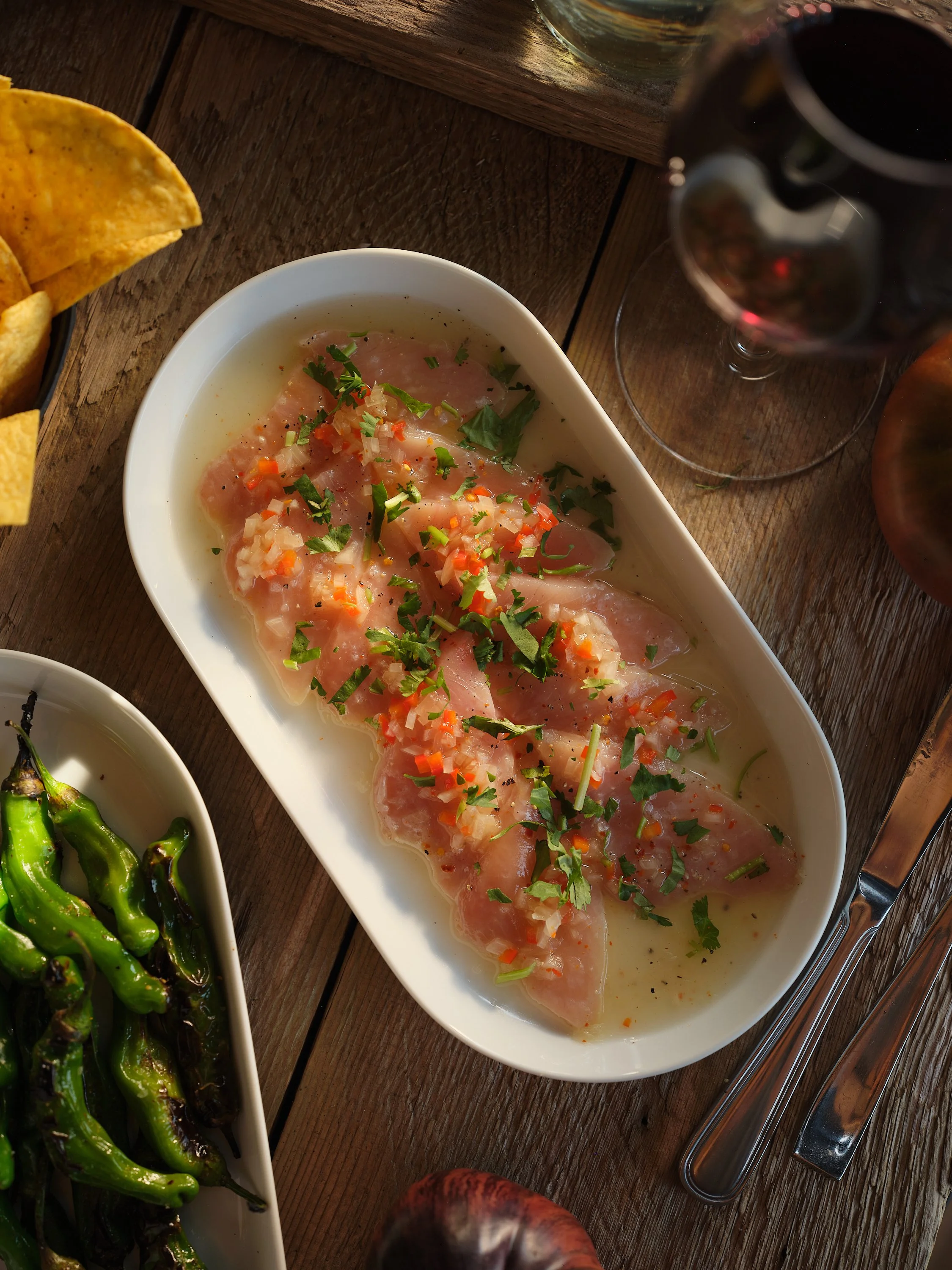 Slices of raw fish garnished with chopped herbs and vegetables served in a white dish on a wooden table, with a glass of red wine and a bowl of roasted green peppers nearby.