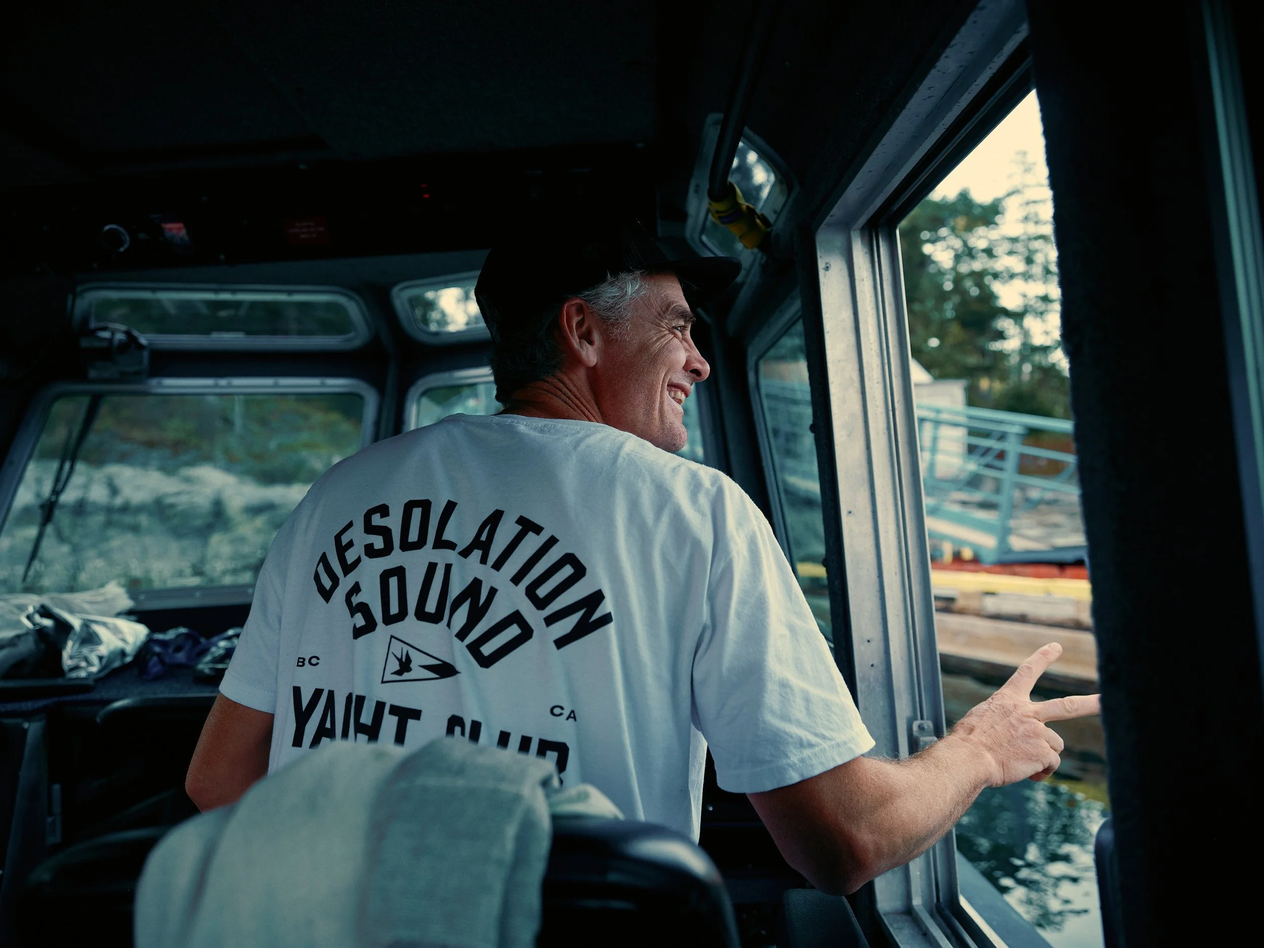 Smiling man with gray hair wearing a black cap and a white T-shirt with black text, sitting inside a boat, pointing at something outside.