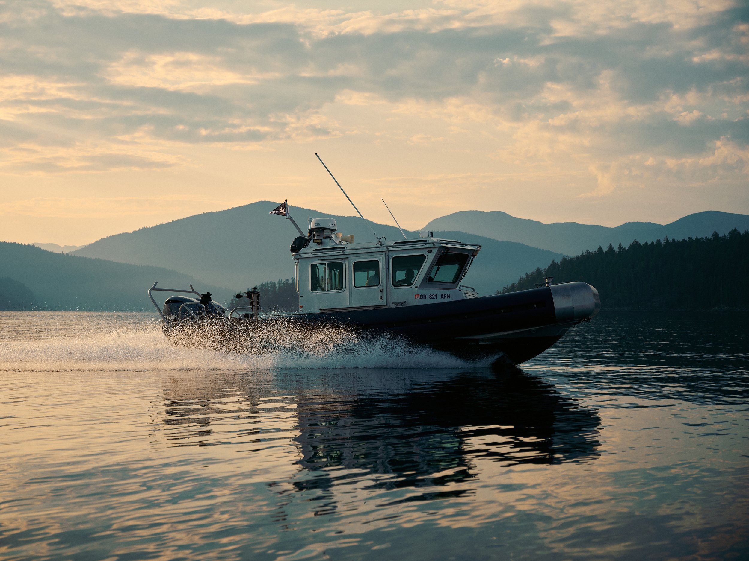 A boat with navigation equipment and antennas on a body of water during sunset, with mountains and a cloudy sky in the background.
