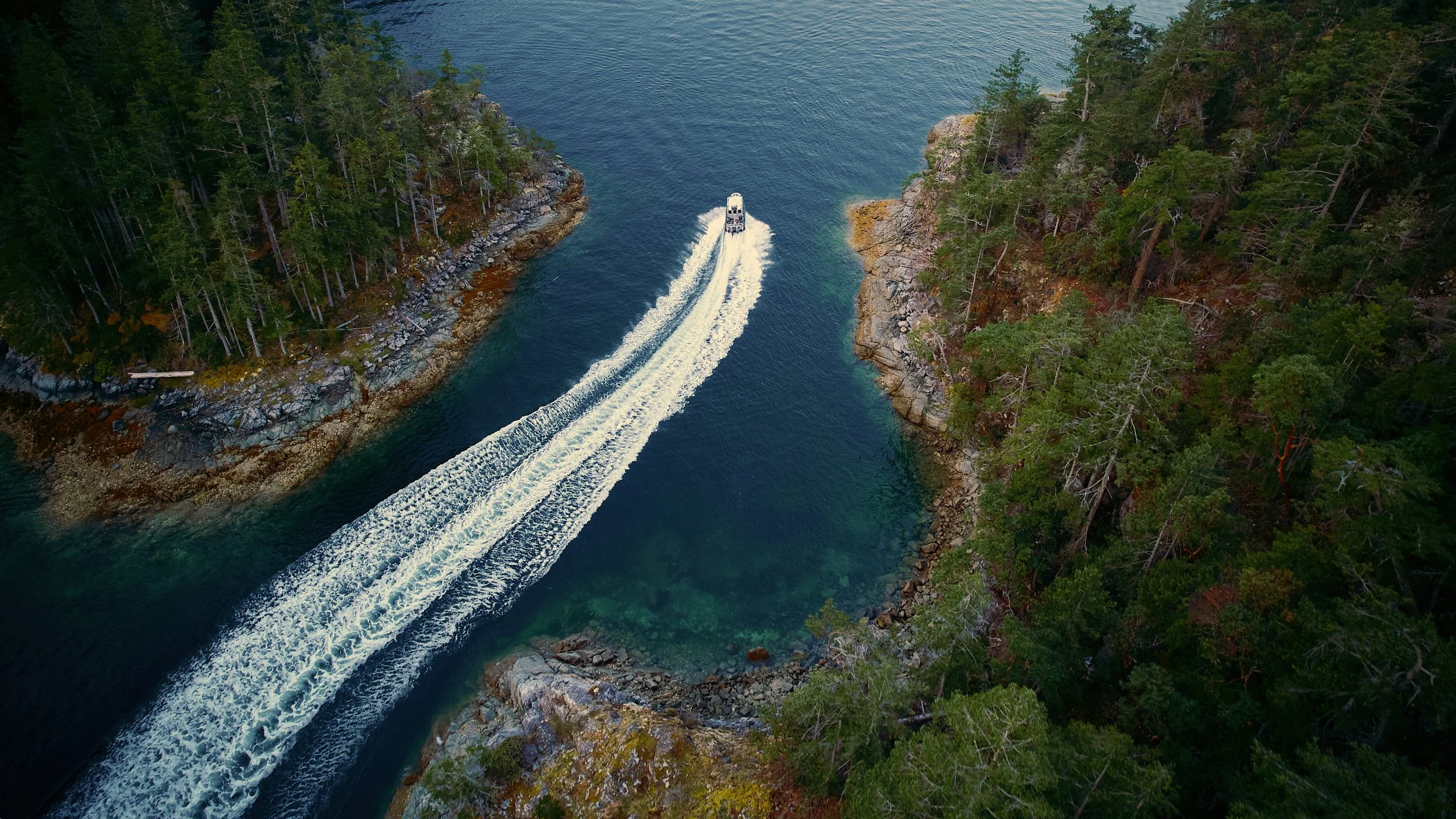 An aerial view of a boat cruising through a narrow, rocky channel between two dense forested islands, creating white wake splashes in the water.