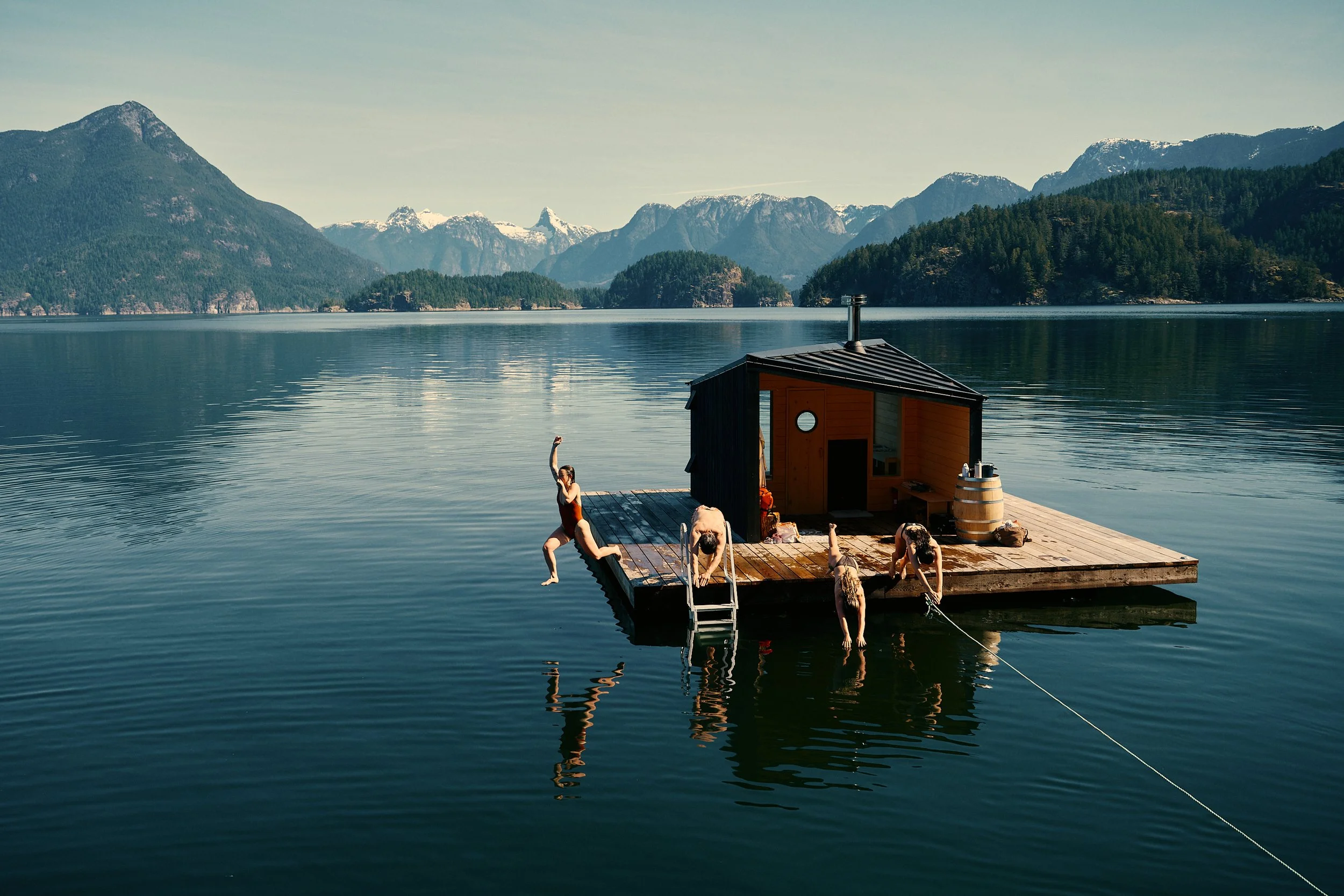 People swimming and relaxing on a wooden dock on calm waters with mountainous landscape in the background.