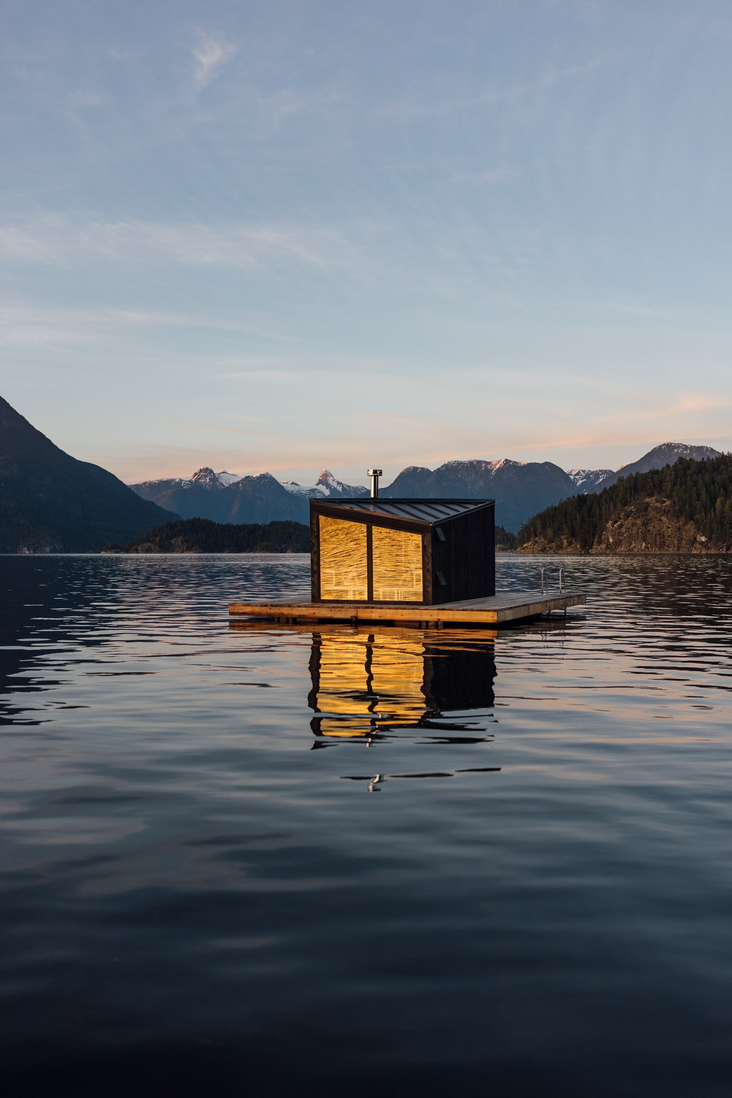 A small illuminated floating sauna sits on a dock in a calm body of water, with mountains and a clear sky in the background during sunset.