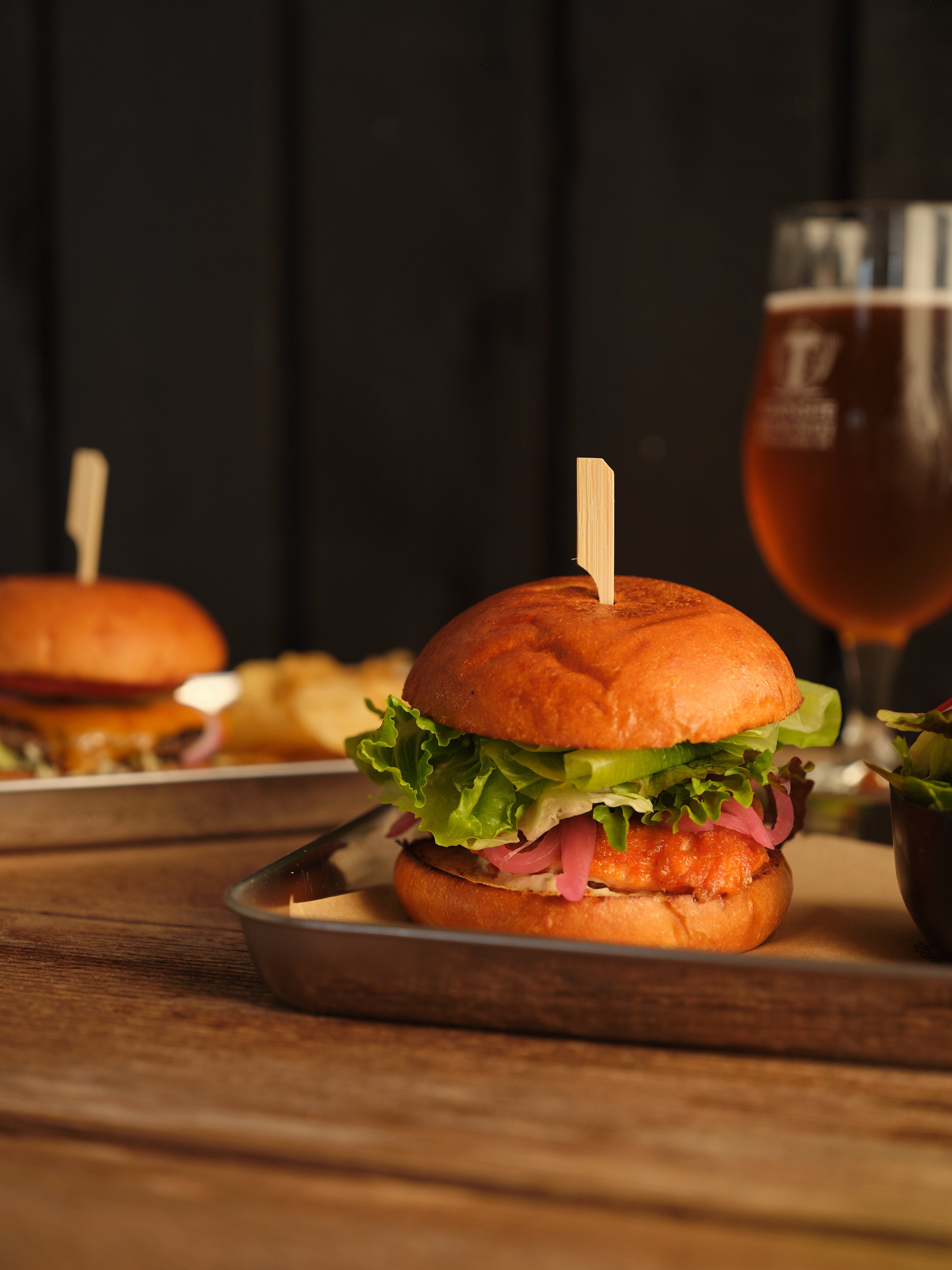 Burger with lettuce, pickles, and pink pickled onions, topped with a bun and a toothpick, placed on a glass plate on a wooden table with another burger and a drink in the background.