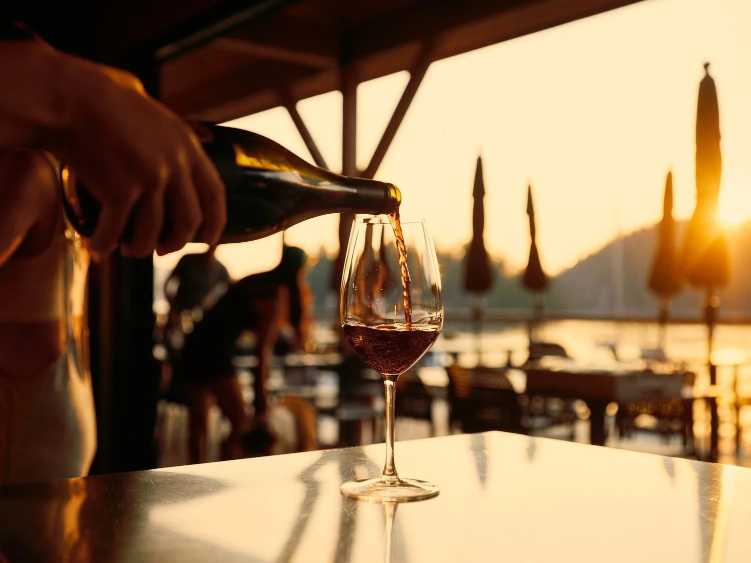 A person pours red wine into a glass on a table at a beachfront bar during sunset.