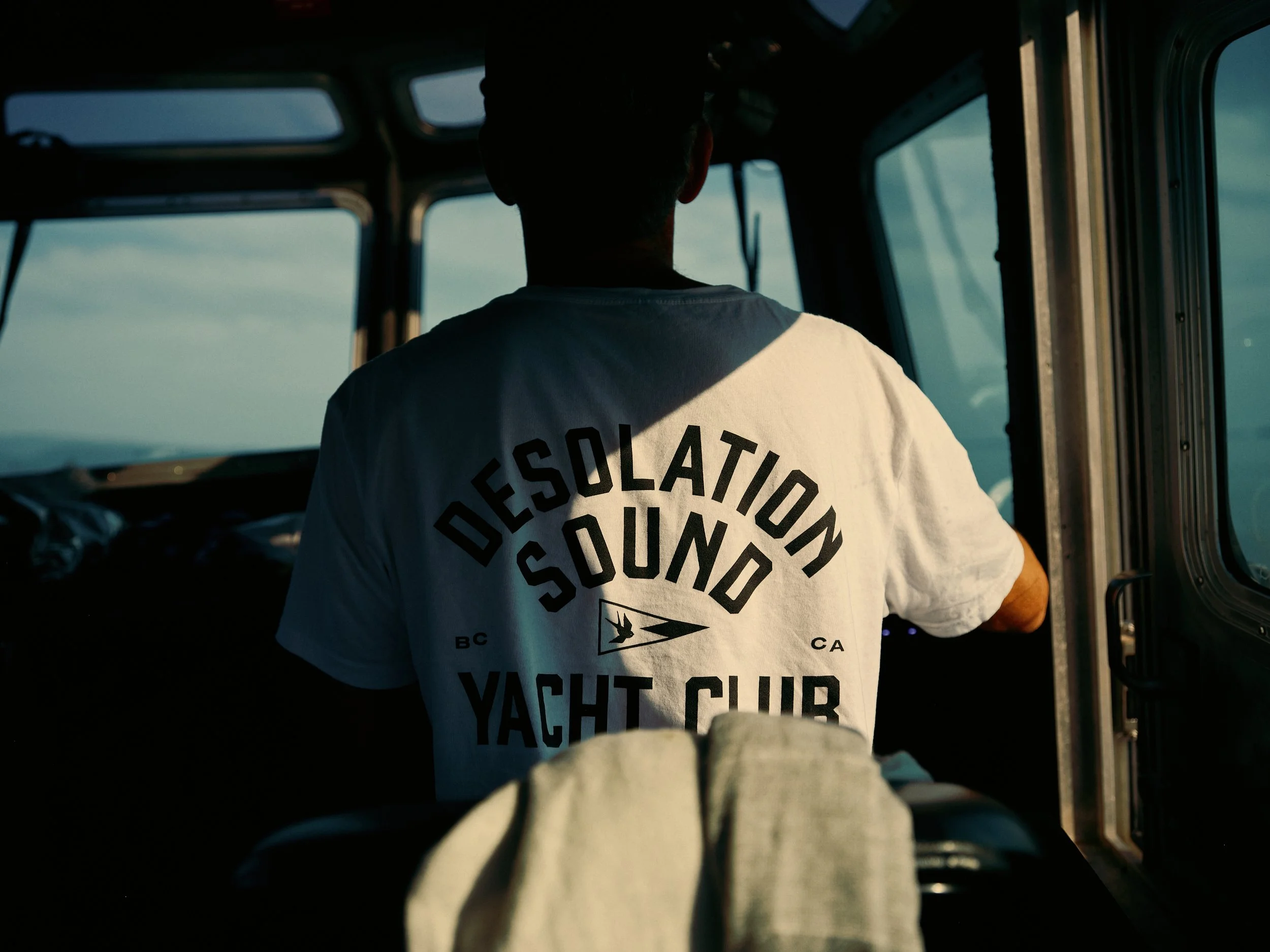 A person seen from behind, wearing a white T-shirt with black text, inside a water taxi with windows showing the sky and landscape outside.