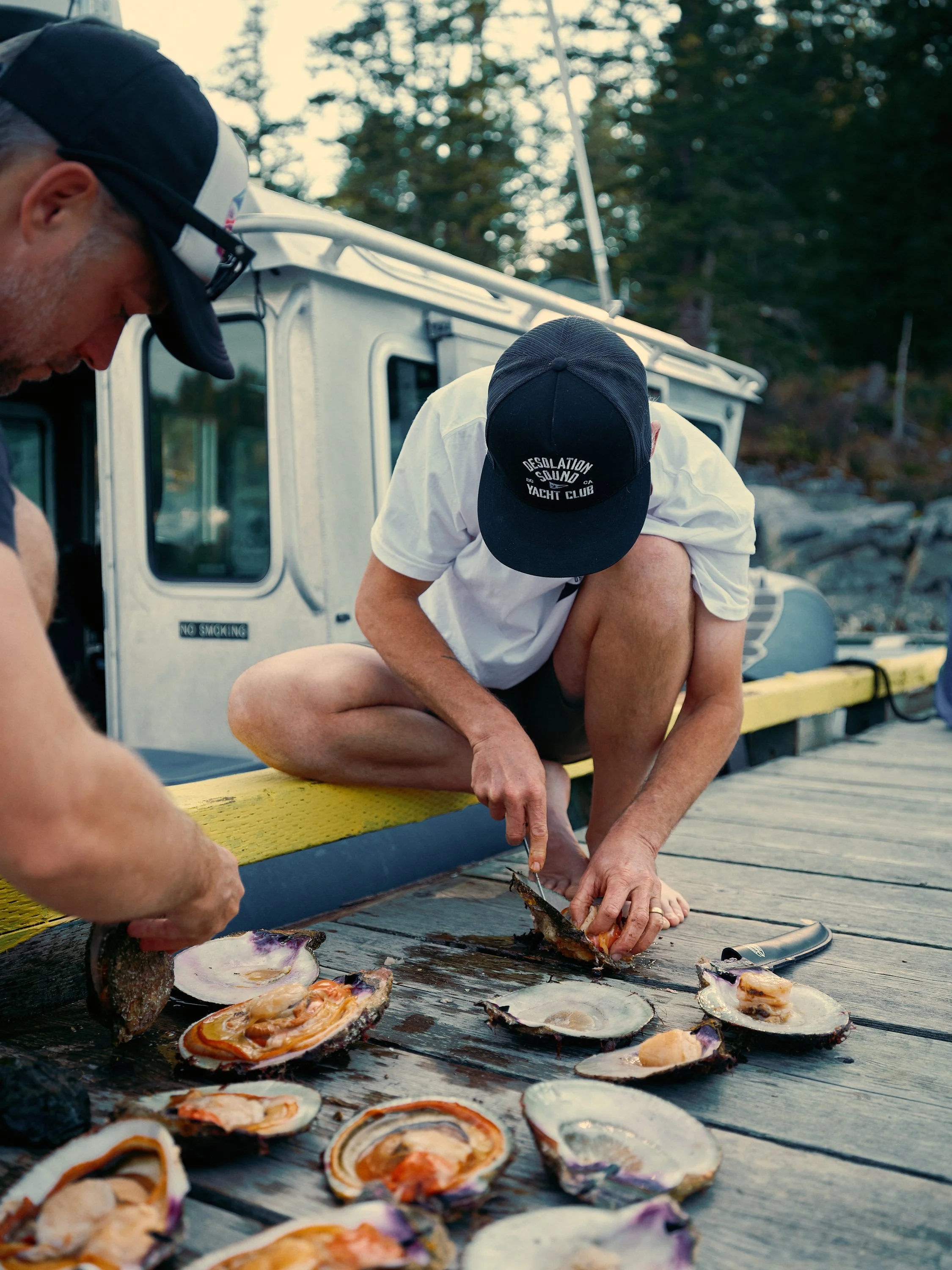 Two men are preparing fresh scallops on a wooden dock with an aluminum boat in the background. One man is squatting and opening the scallops, while the other man stands nearby. The scene takes place outdoors with trees in the background.
