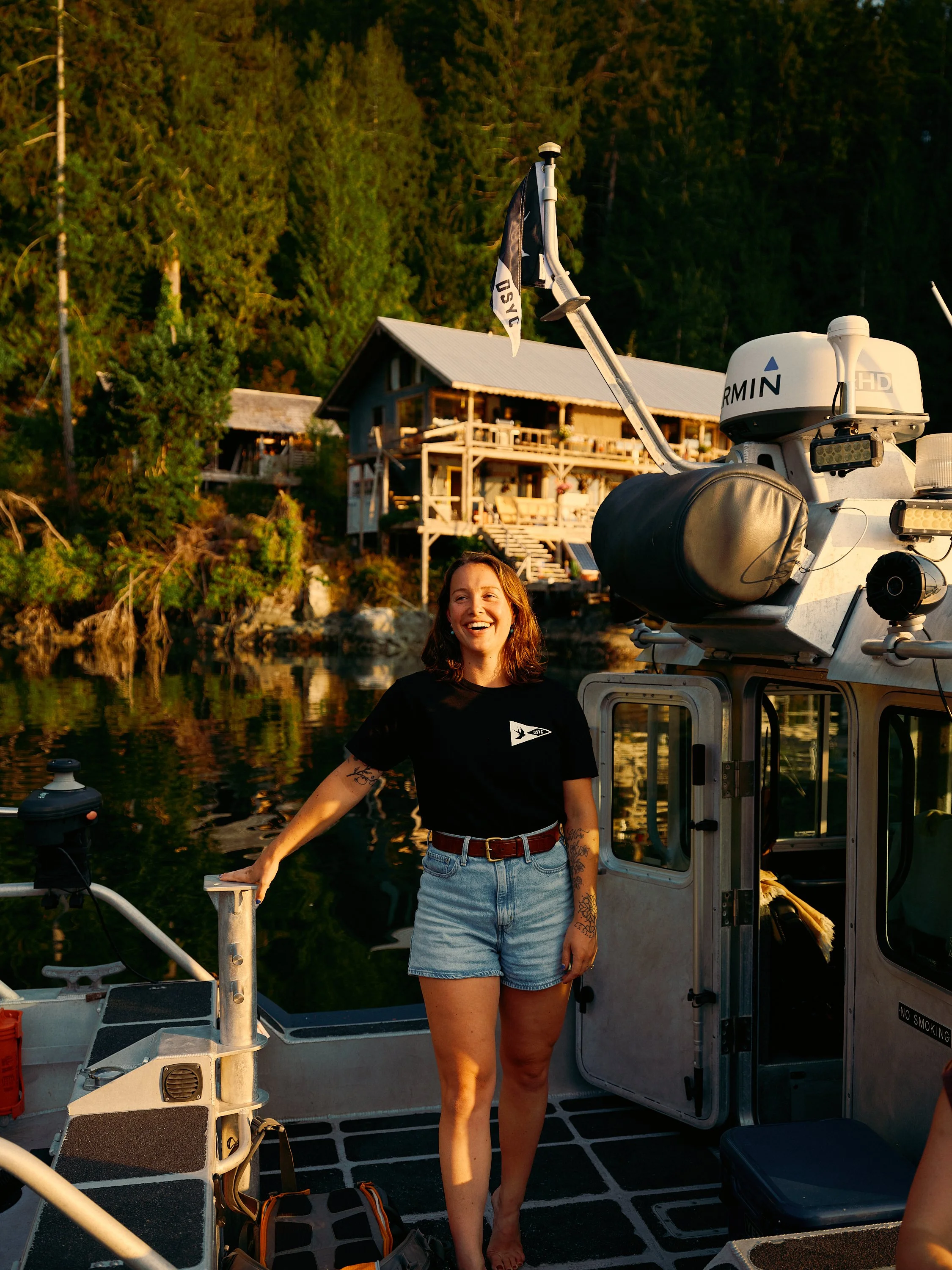 A woman standing on a boat's deck, smiling, with a Garmin marine GPS and radar equipment nearby, and a wooden house on stilts surrounded by trees in the background.