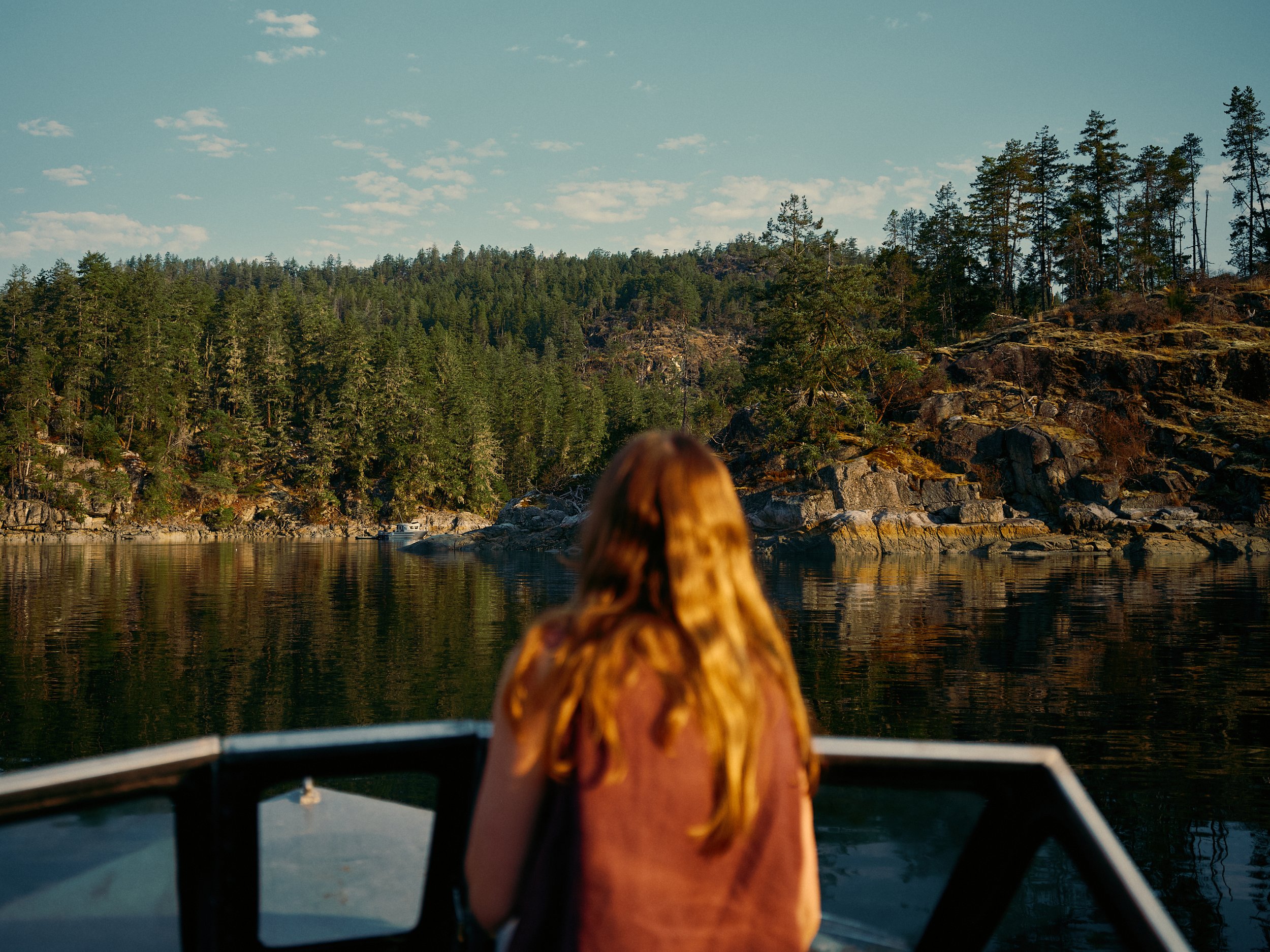 A person with long hair sitting at the front of a boat on calm waters, gazing out at a forested landscape with a hill and some clouds in the sky.