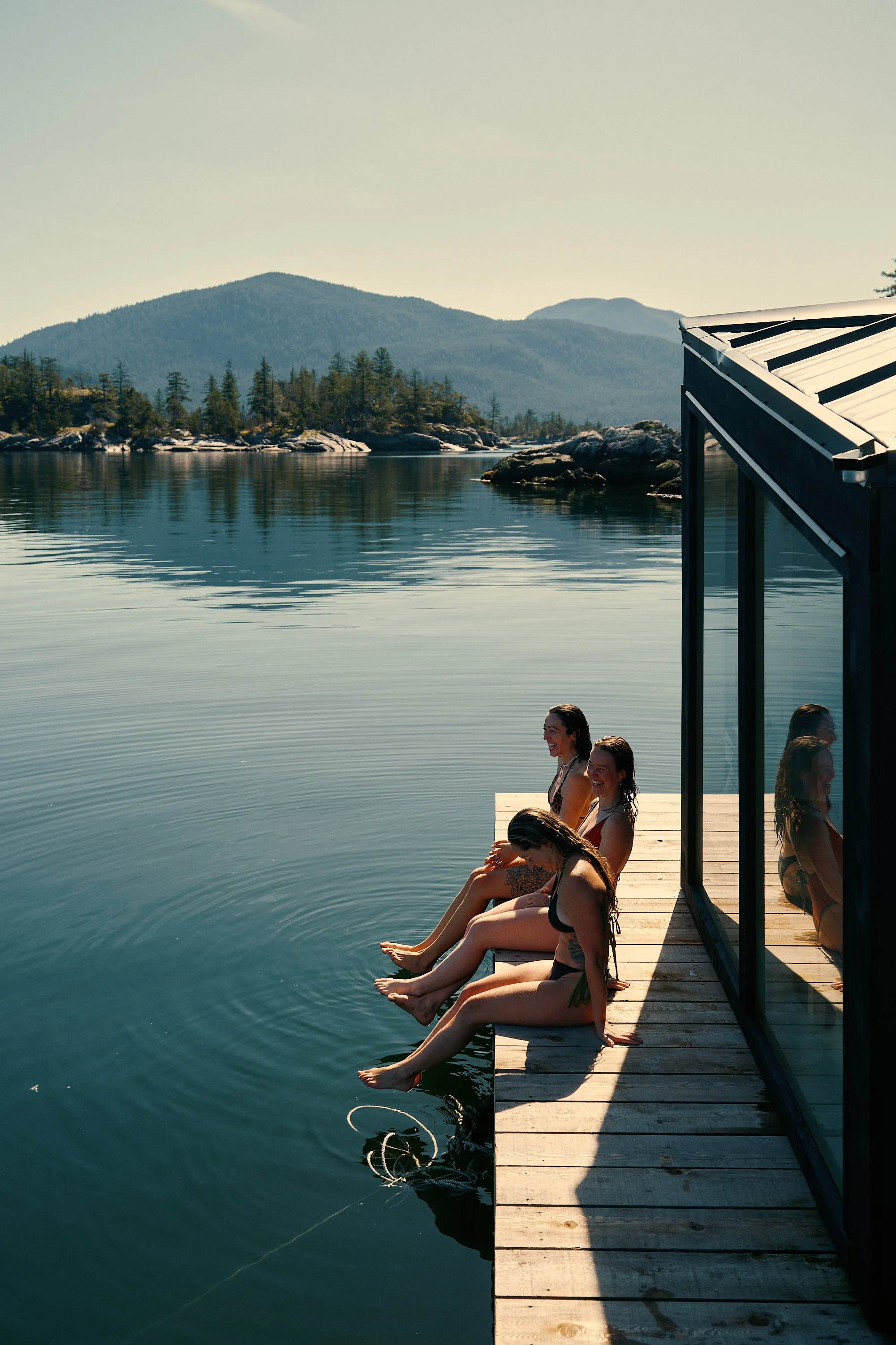 People sitting on the dock of a floating sauna with their feet hanging in the water, during daytime, with mountains and trees in the background, and a building structure on the right.