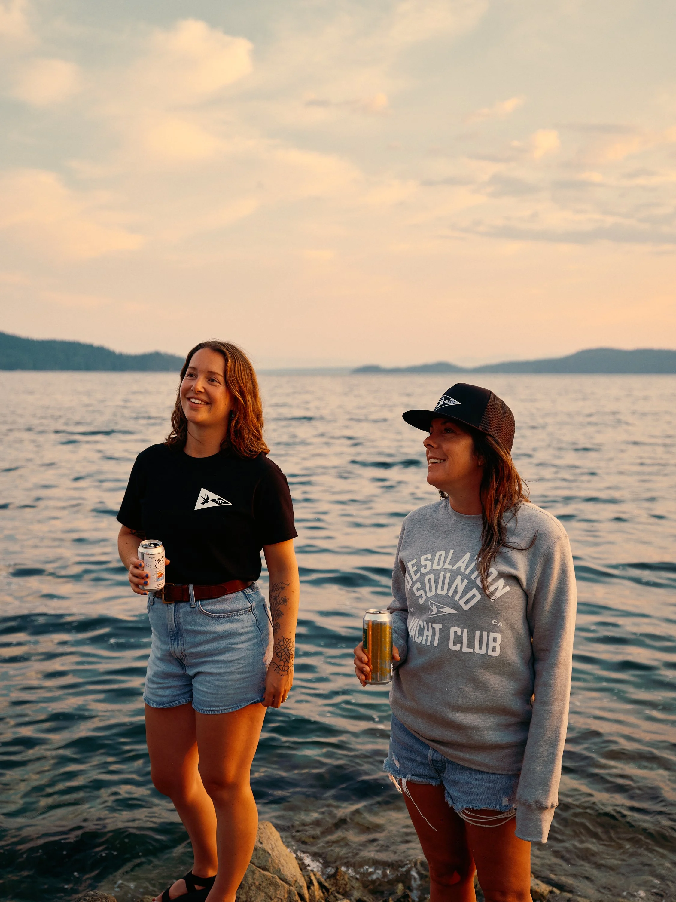 Two women standing on rocks by the water during sunset, smiling and holding beverage cans, with a scenic lake and distant mountains in the background.