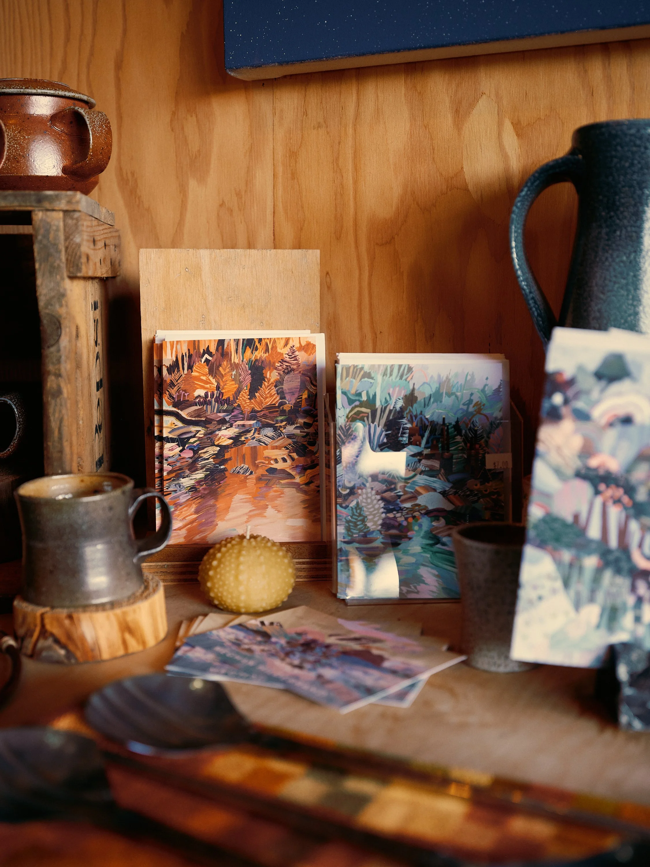 Display of colorful postcards or prints with abstract and nature-inspired designs arranged on a wooden surface, with ceramic cups and a yellow spiky object in the foreground.
