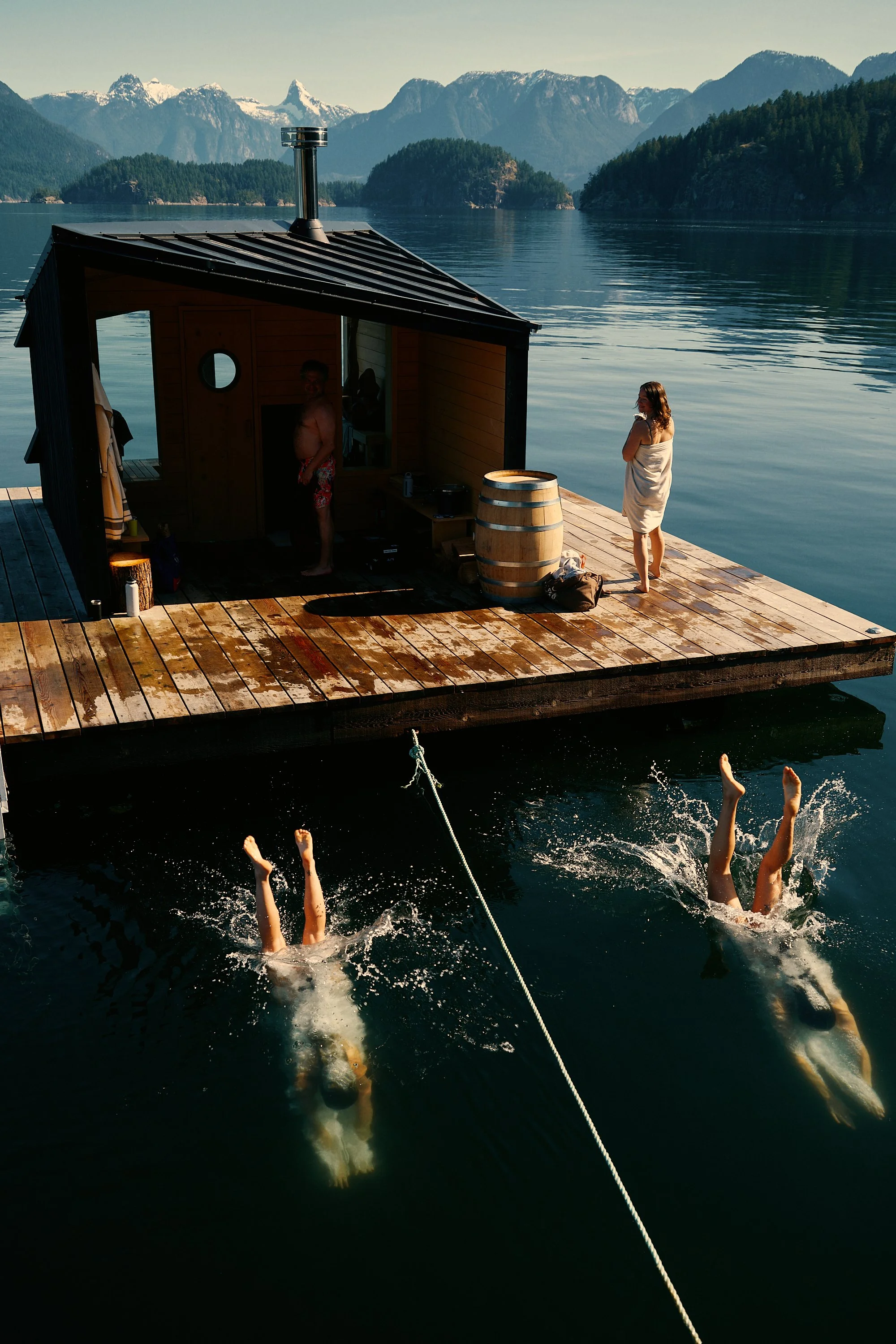 A person swimming underwater near a dock, and two people standing on the dock, with mountain and forest scenery in the background.