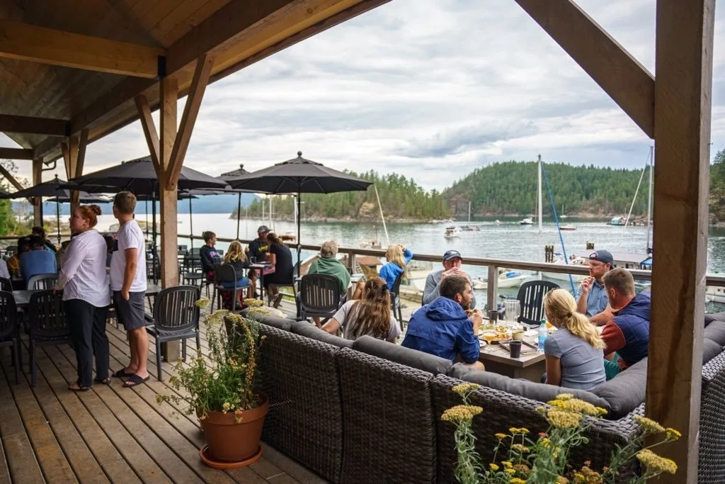 People dining and socializing on a covered wooden deck overlooking a lake with boats and forested hills in the background.