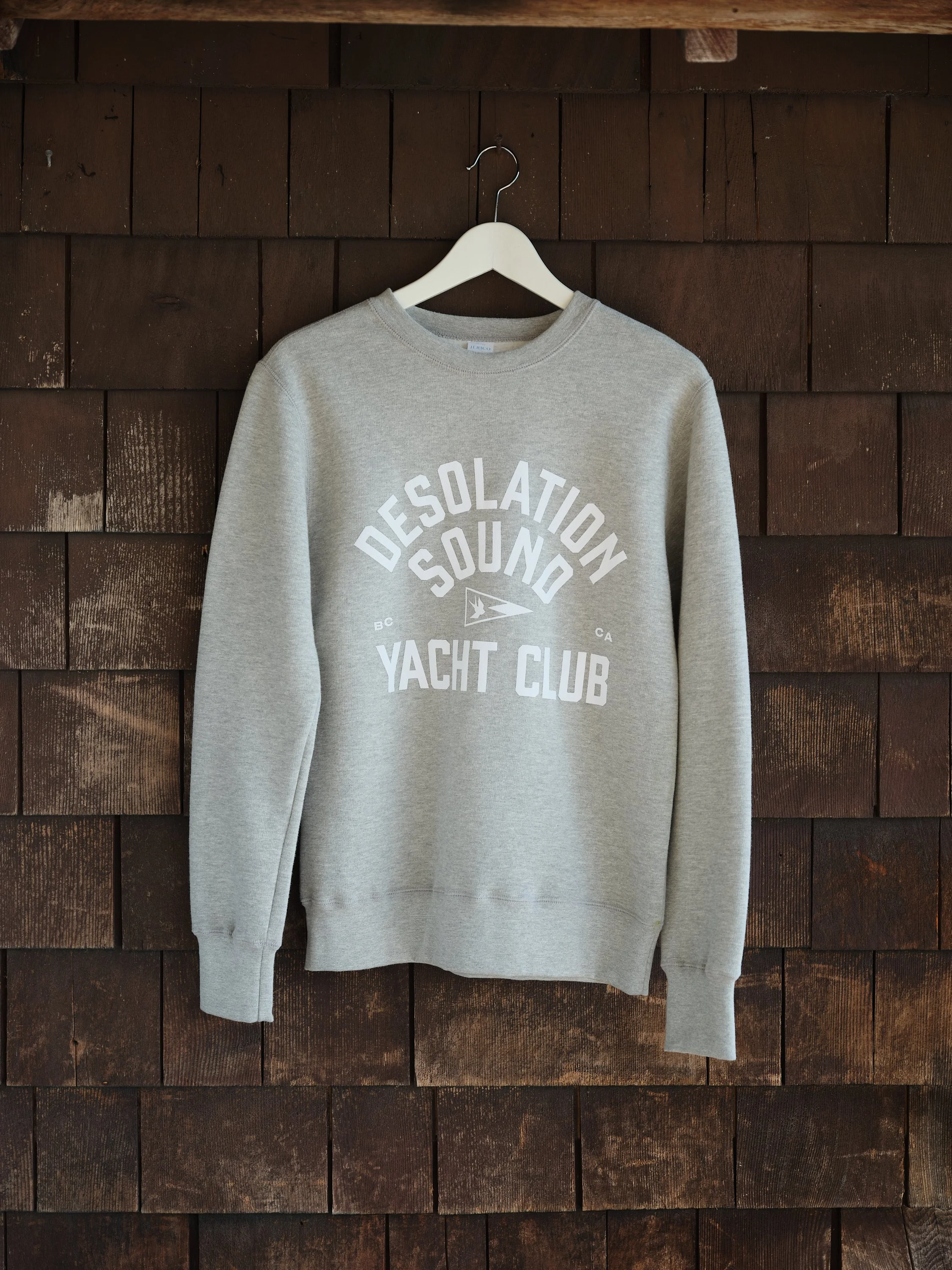 A gray sweatshirt with the words 'Desolation Sound' and 'Yacht Club' printed on the front hanging on a white hanger against a wooden wall background.