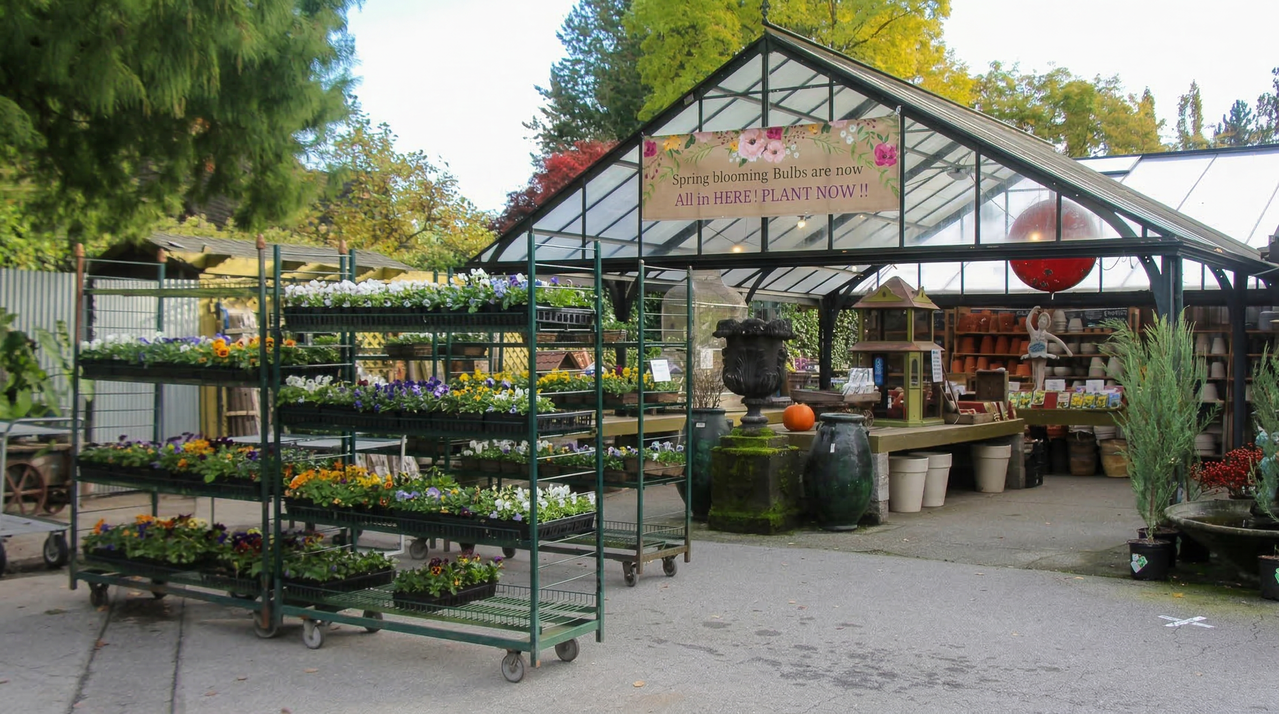 A garden center with colorful flowers on metal carts and shelves, a greenhouse with a sign that reads 'Spring blooming bulbs are now all in here! Plant now!', and trees with green and fall foliage in the background.