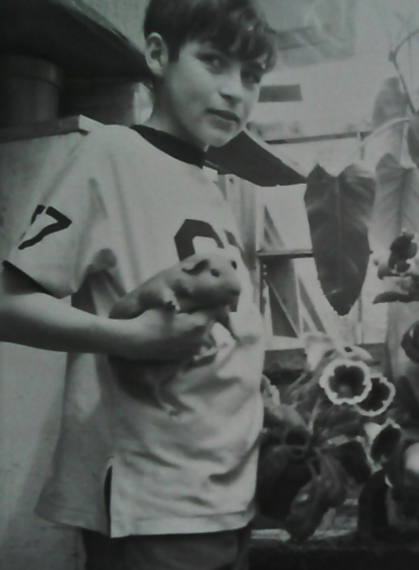 Young man with curly hair wearing a sports jersey holding a small pig in his hands, standing indoors by plants and other objects.