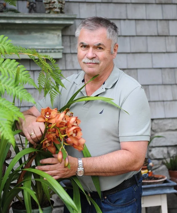 A middle-aged man with gray hair and a mustache standing outdoors in front of a grey wooden wall, holding a potted orange orchid plant, with green plants around him.