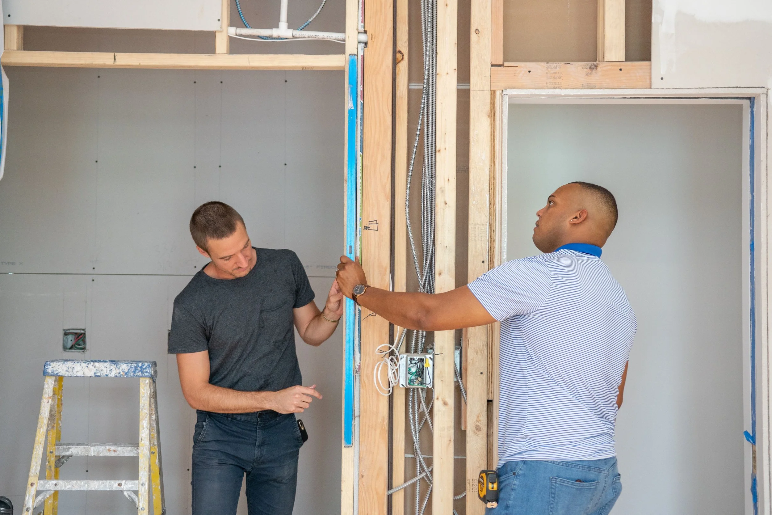 Two men working on electrical wiring inside a construction site, with exposed wooden framing and visible wires.
