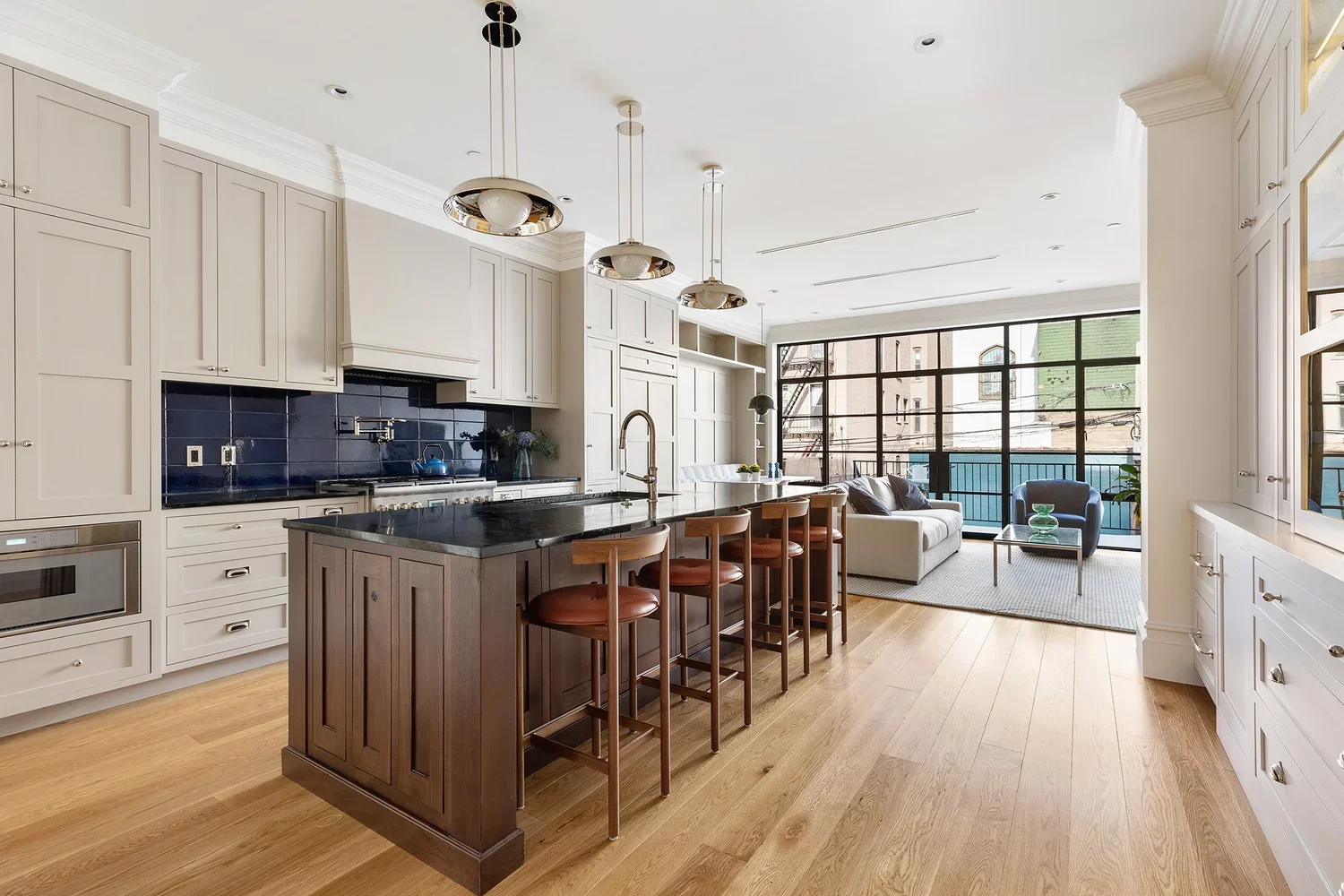 Open-concept living room and kitchen with white cabinetry, black countertops, wooden stools at a kitchen island, and large windows in the living area with seating and decor.