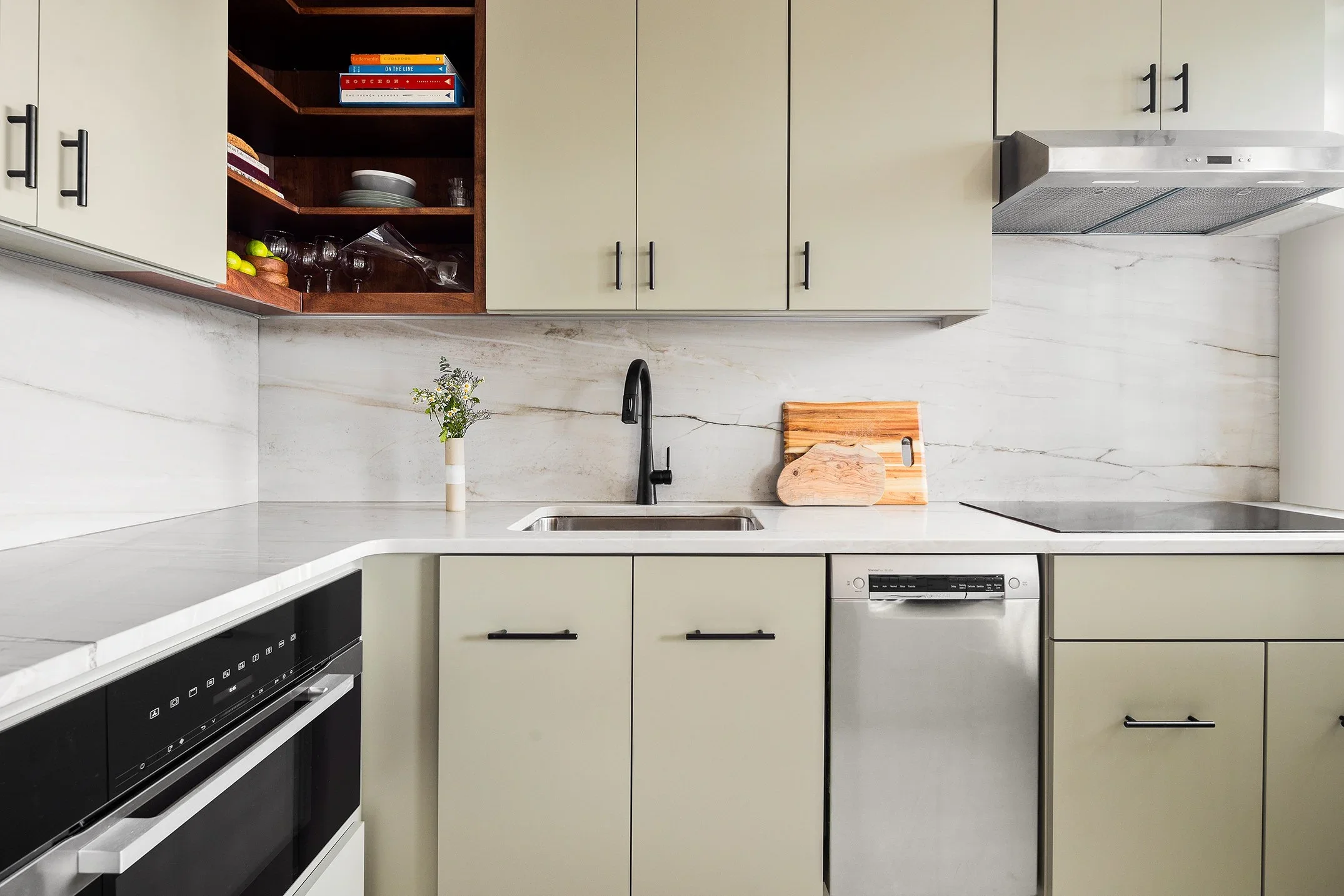 Modern kitchen with light green cabinets, marble countertop, black faucet, open wooden shelves with books and dishes, vase with flowers, cutting board, and built-in oven.