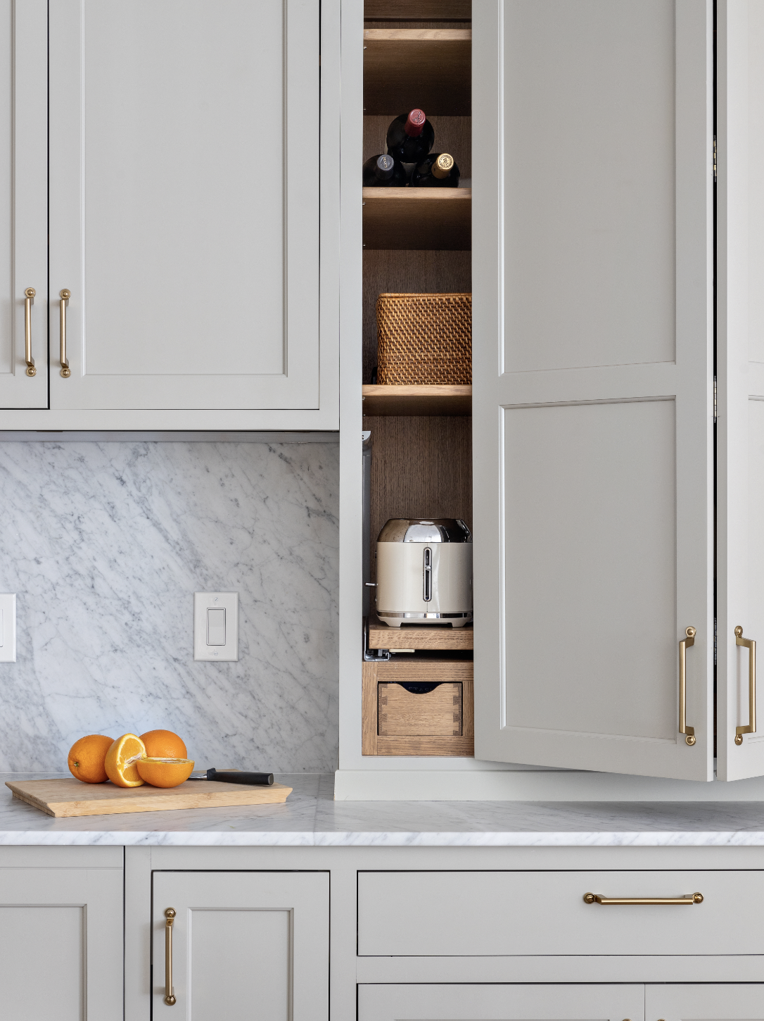 Kitchen cabinet with wine bottles, a woven basket, a silver toaster, oranges, and a marble countertop.