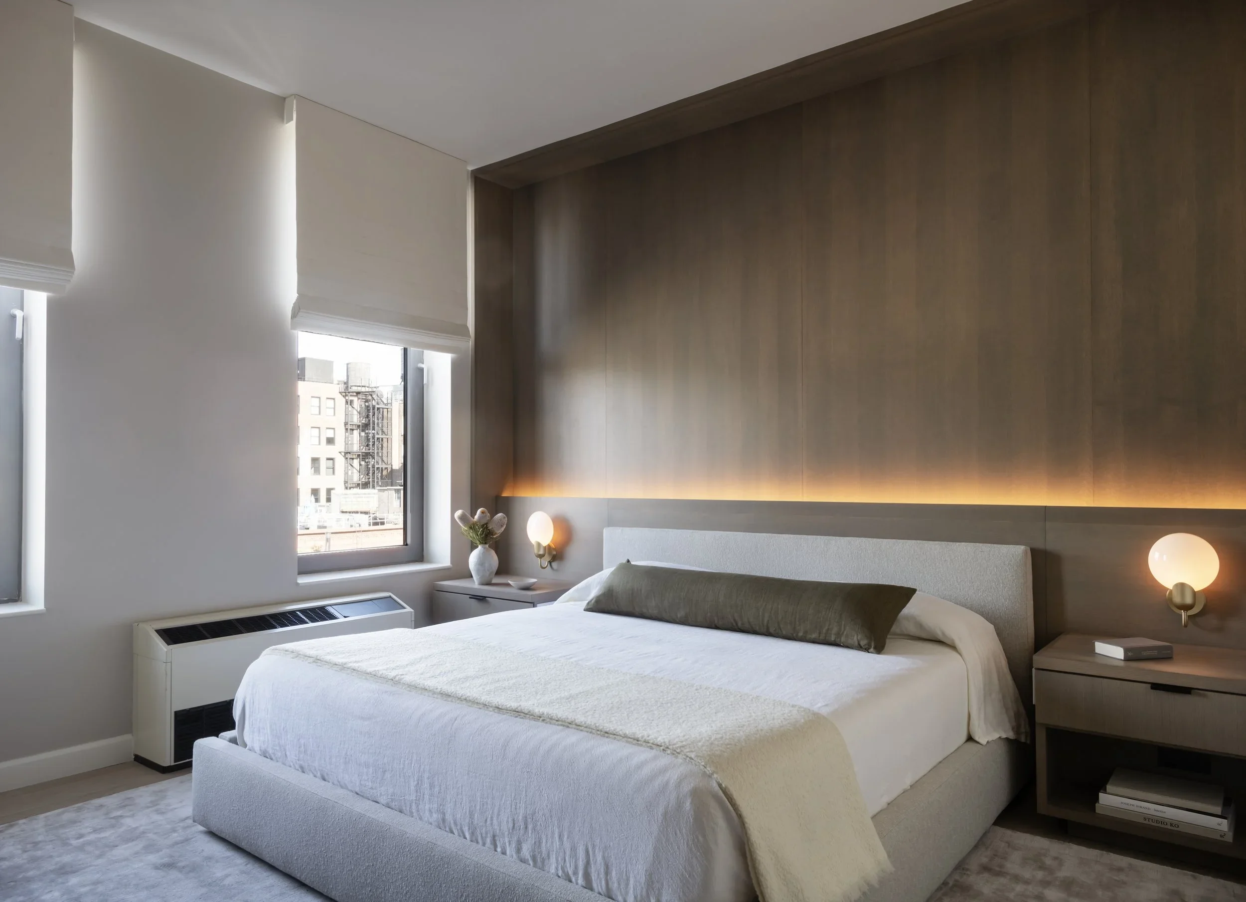 Modern bedroom with white bed, beige pillows, nightstand, wall lamps, wooden wall panel, window with city view, vase with flowers, and neutral color scheme.
