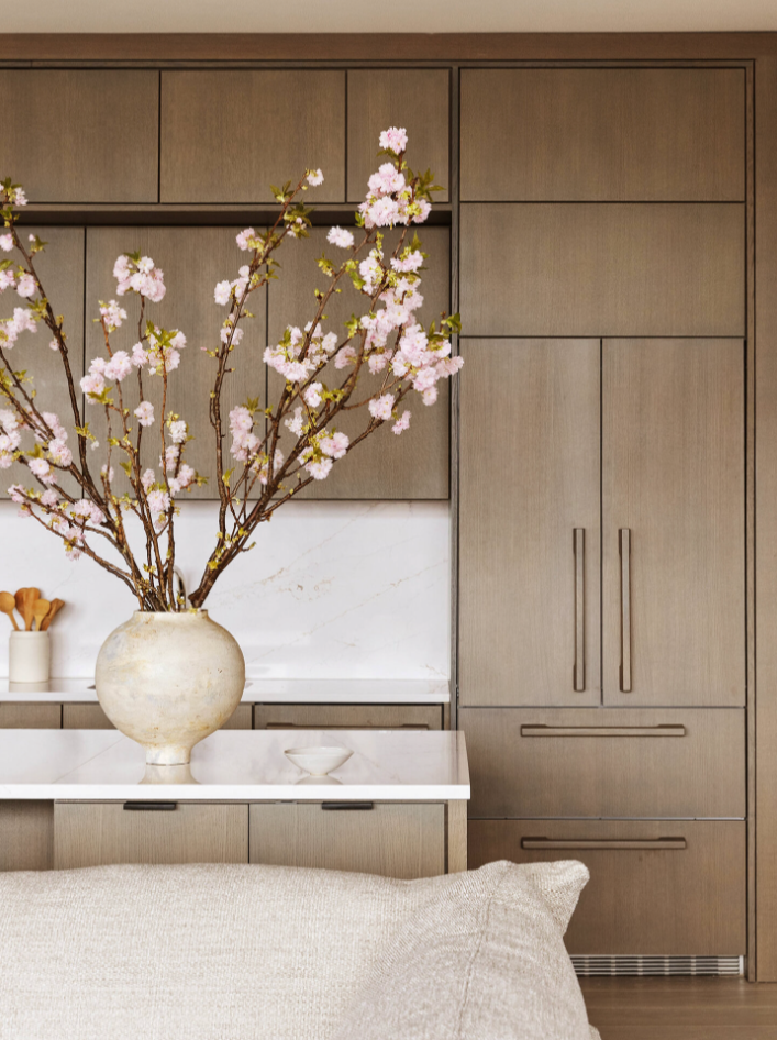 Interior of a modern kitchen with wooden cabinets, a white marble backsplash, and a white countertop. A large ceramic vase with pink cherry blossom branches is on the counter. Part of a beige sofa is visible in the foreground.