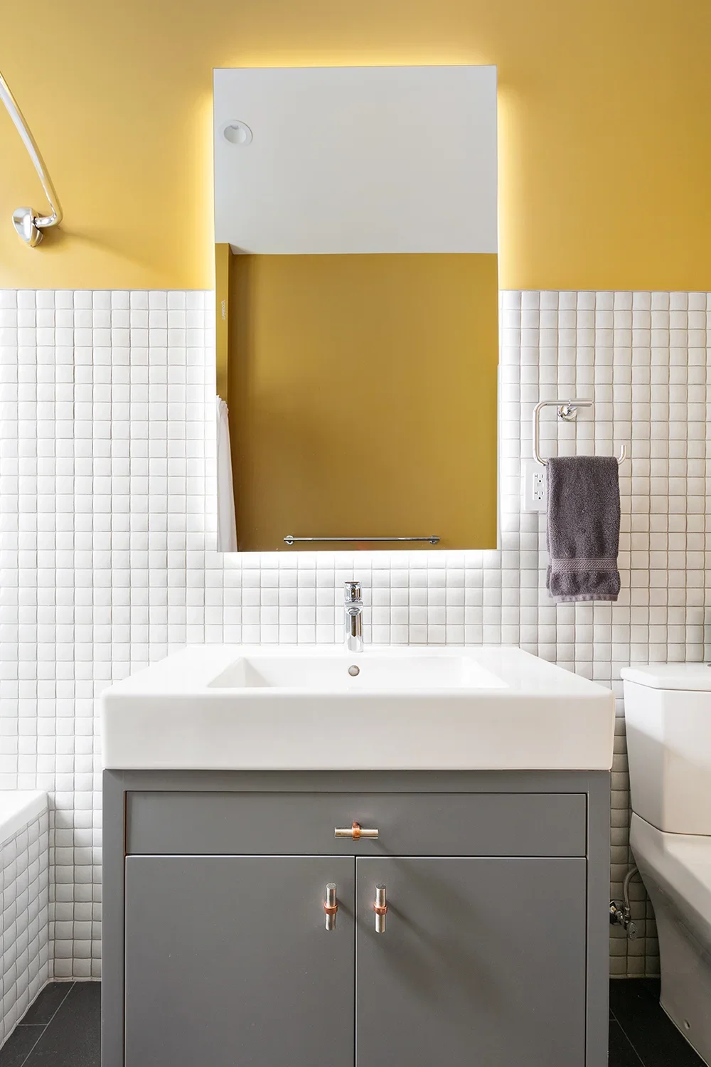 Modern bathroom with white tiled walls, yellow upper walls, and a large mirror above a grey vanity with a white sink. A towel is hanging on the right, and part of a toilet is visible on the right side.