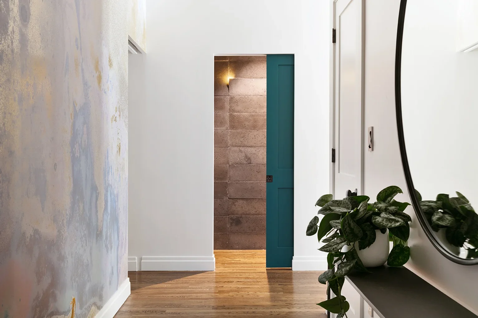 Indoor hallway with wood flooring, a potted plant on a wall shelf, a mirror, and a partially open teal door leading to a room with a textured brown wall and a wall sconce