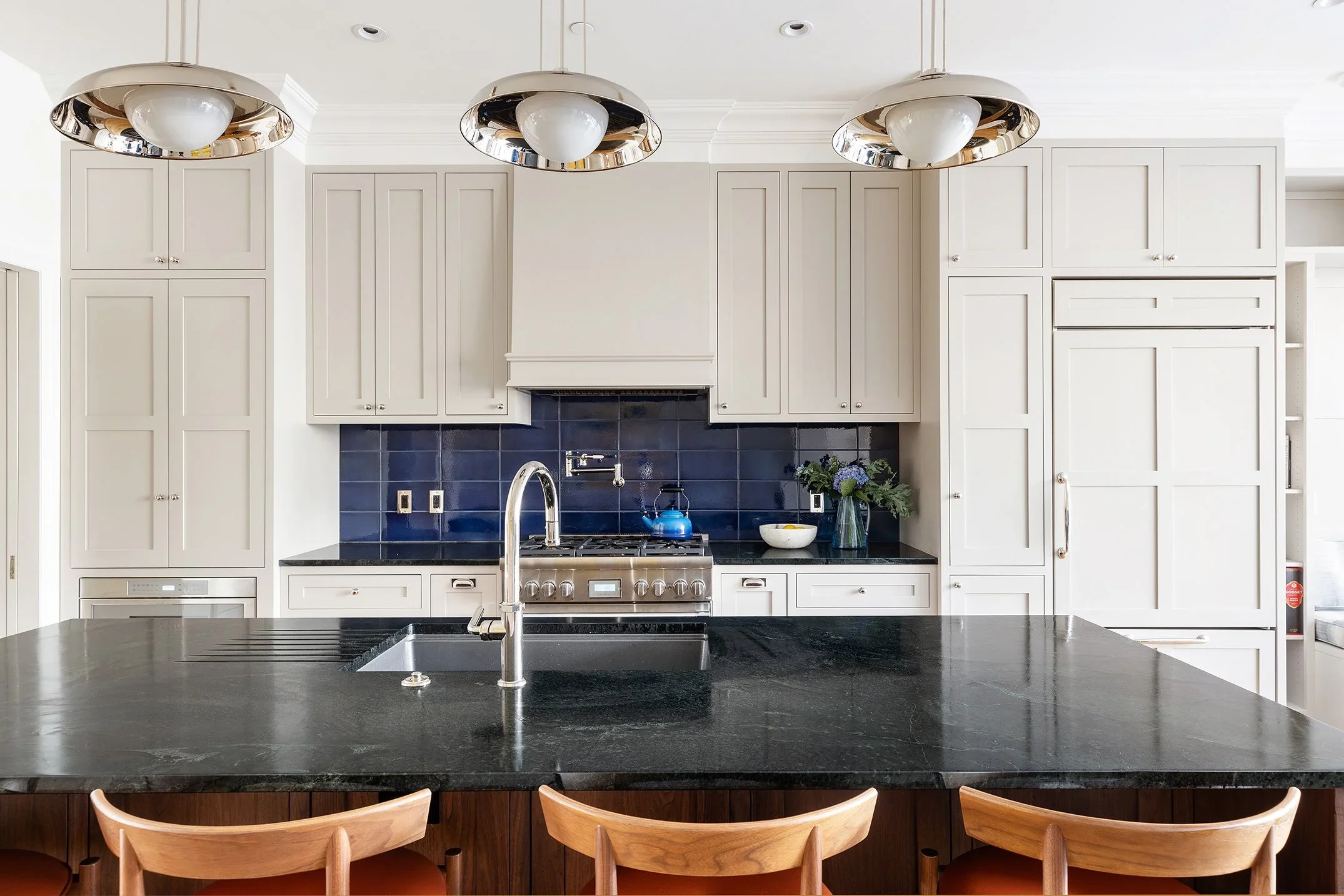 Modern kitchen with white cabinets, black countertops, stainless steel stove, blue backsplash, and a black island with a sink and four wooden chairs.