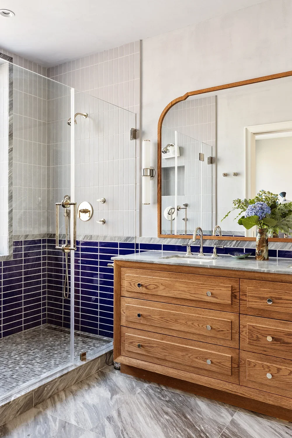 Modern bathroom with a wooden vanity, marble countertop, large mirror, glass shower with blue and white tiles, and a bouquet of flowers.