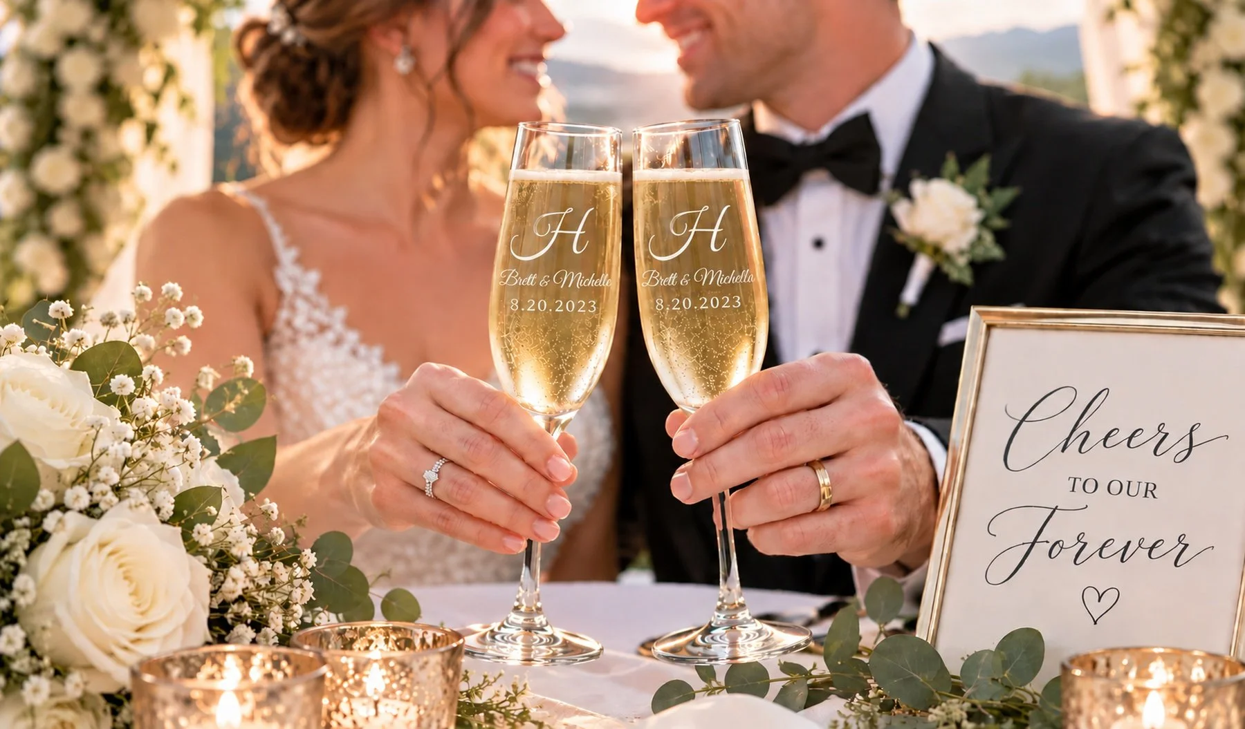 A bride and groom at their wedding celebration, holding glasses of champagne. The glasses are engraved with the initials 'H,' the names Brett and Michelle, and the date 8.20.2023. There are floral arrangements and a sign that says 'Cheers to our forever.'
